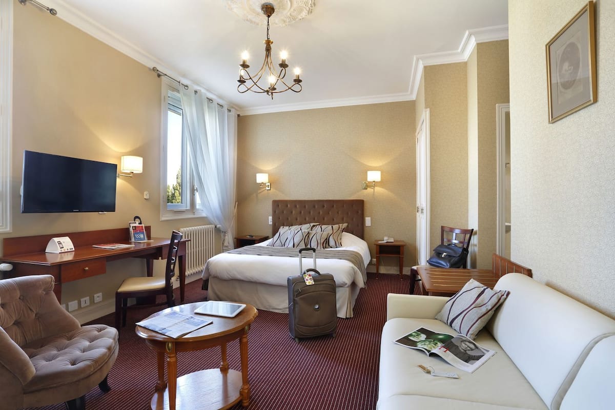 Habitación de hotel con cama, escritorio, sillón, sofá, televisor y candelabro.