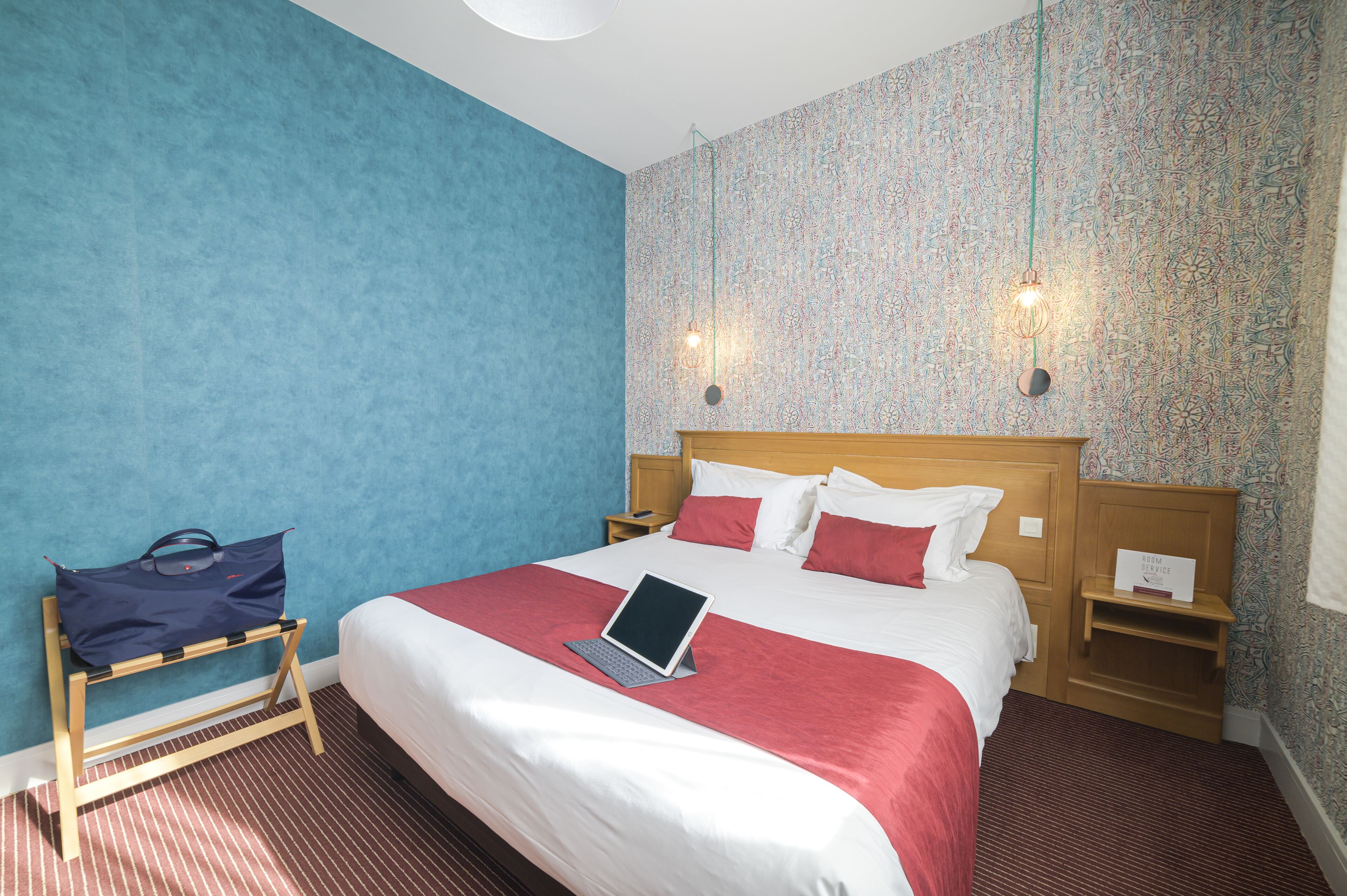 A hotel room with a bed, a laptop on a bedside table, a blue wall, a patterned wallpaper, and a wooden nightstand.