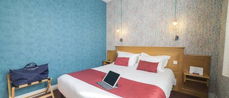 Una habitación de hotel con una cama, un ordenador portátil sobre una mesita de noche, una pared azul, un papel pintado con diseño y un mueble de noche de madera.