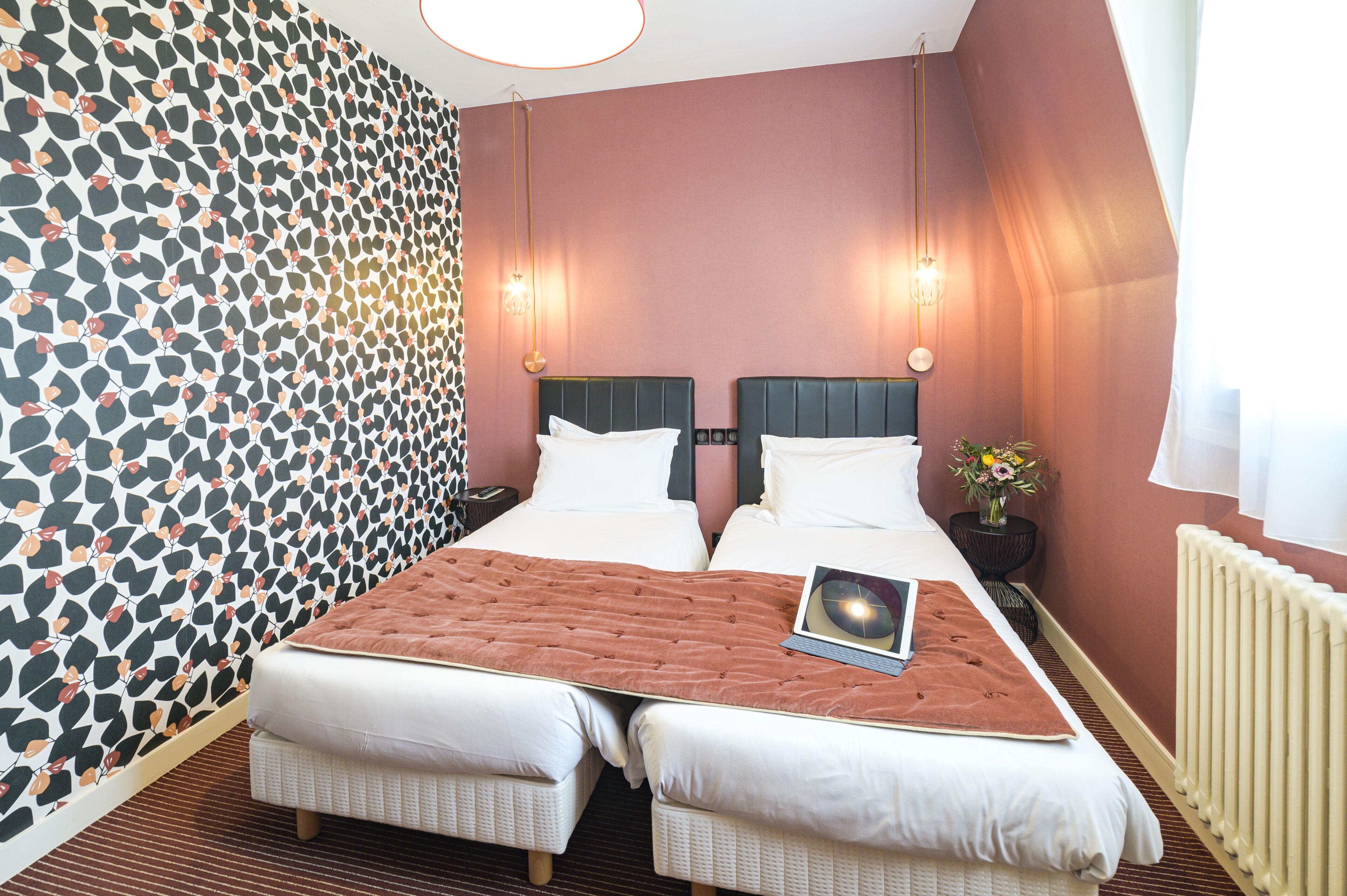 A double bed with a laptop, two bedside tables, a vase with flowers, and a patterned wallpaper.