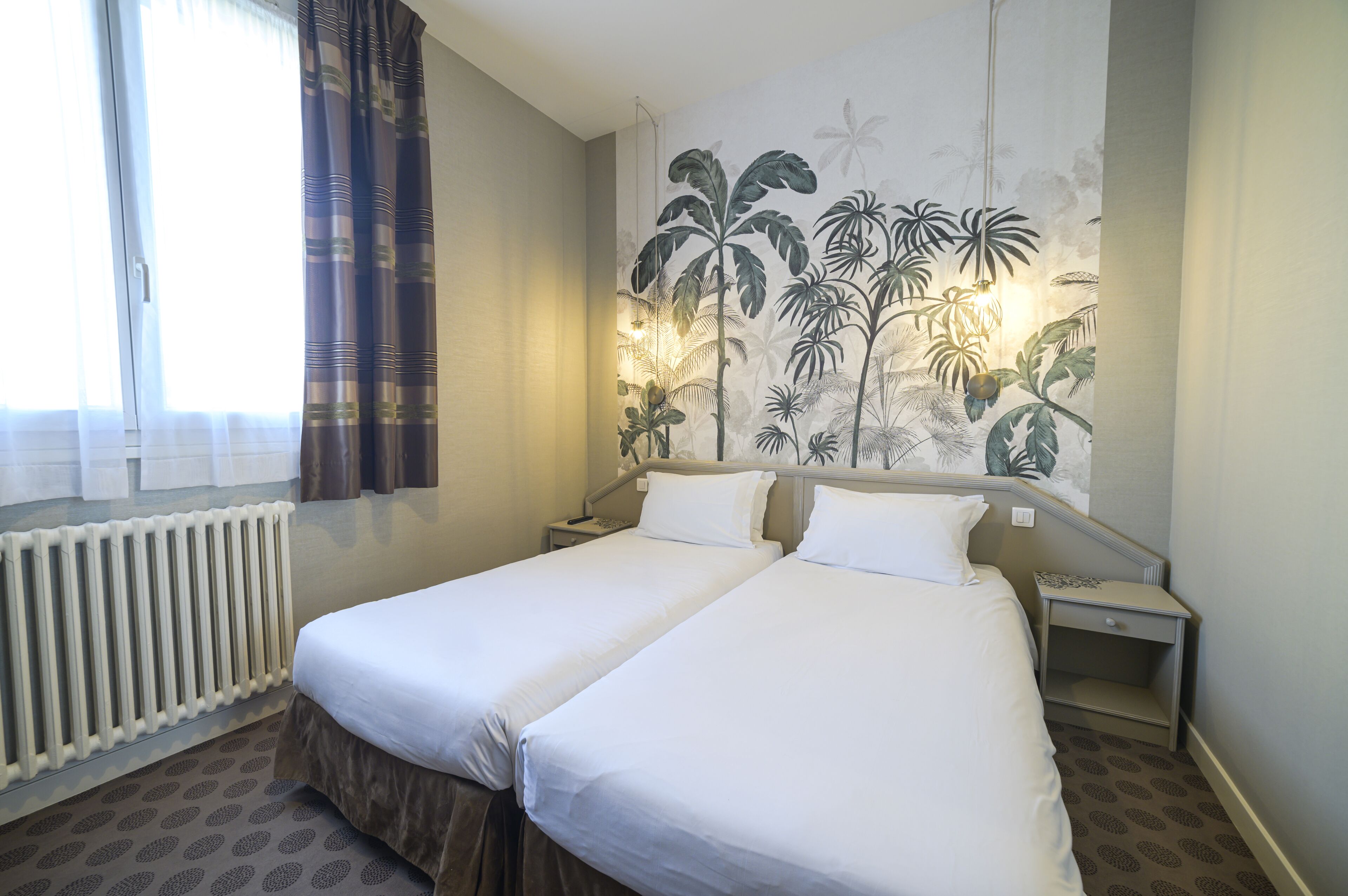 A hotel room with two single beds, a radiator, a window with curtains, and a wall mural of tropical plants.