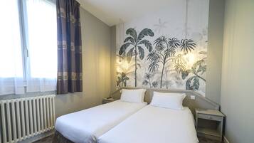 A hotel room with two single beds, a radiator, a window with curtains, and a wall mural of tropical plants.