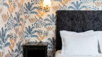 A hotel room with a patterned wallpaper, a black nightstand, a white bed with pillows, and a wall-mounted lamp.
