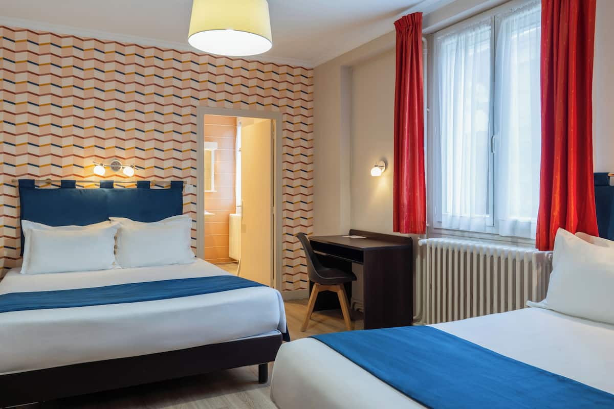 Habitación de hotel con dos camas, un escritorio y una silla. Se ve una ventana con cortinas rojas y un radiador.