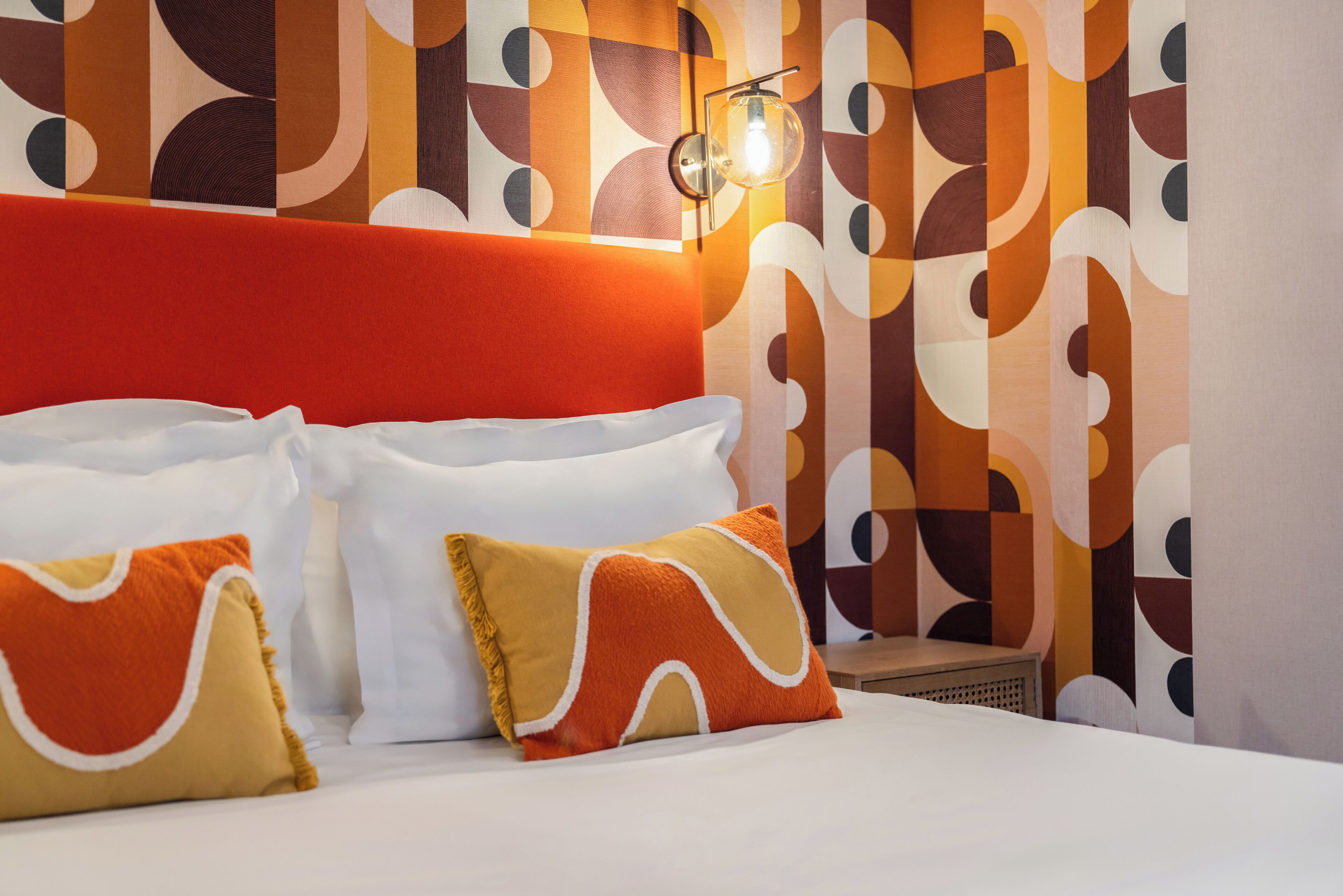 Habitación de hotel con diseño moderno, que incluye una cama grande con almohadas blancas y anaranjadas, un cabecero rojo y una pared con estampado.