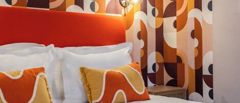 A hotel room with a modern design, featuring a large bed with white and orange pillows, a red headboard, and a patterned wall.