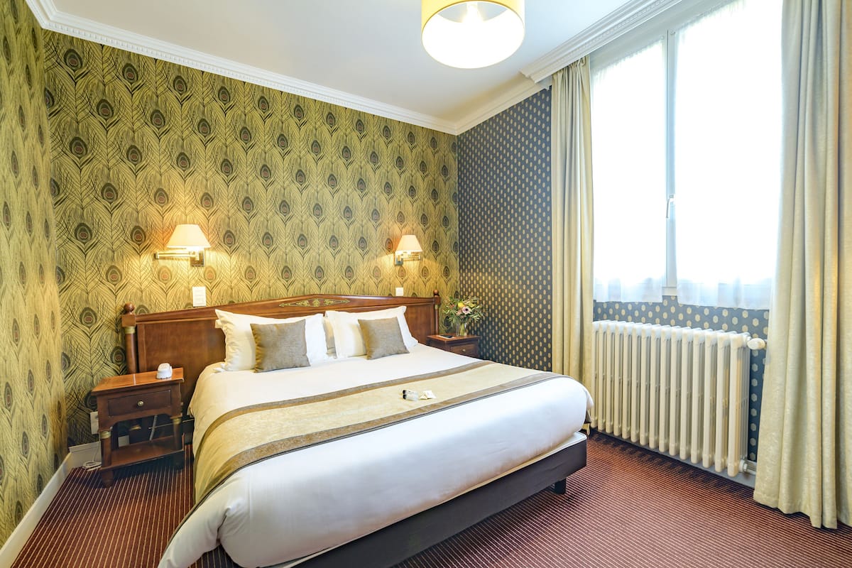 Habitación de hotel con una cama grande, dos mesitas de noche, un radiador y una ventana con cortinas.
