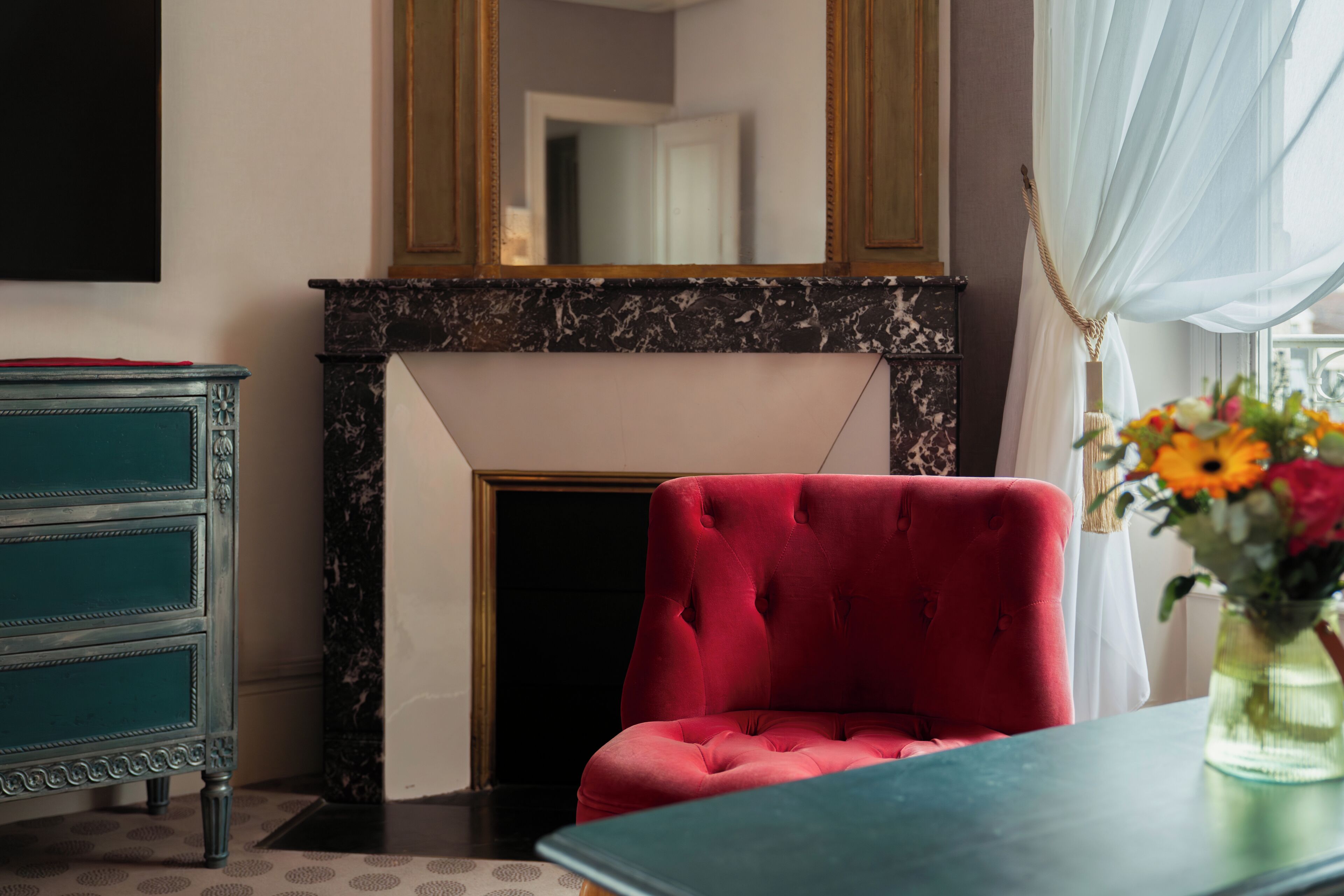 A room with a red armchair, a teal dresser, and a fireplace with a marble surround.