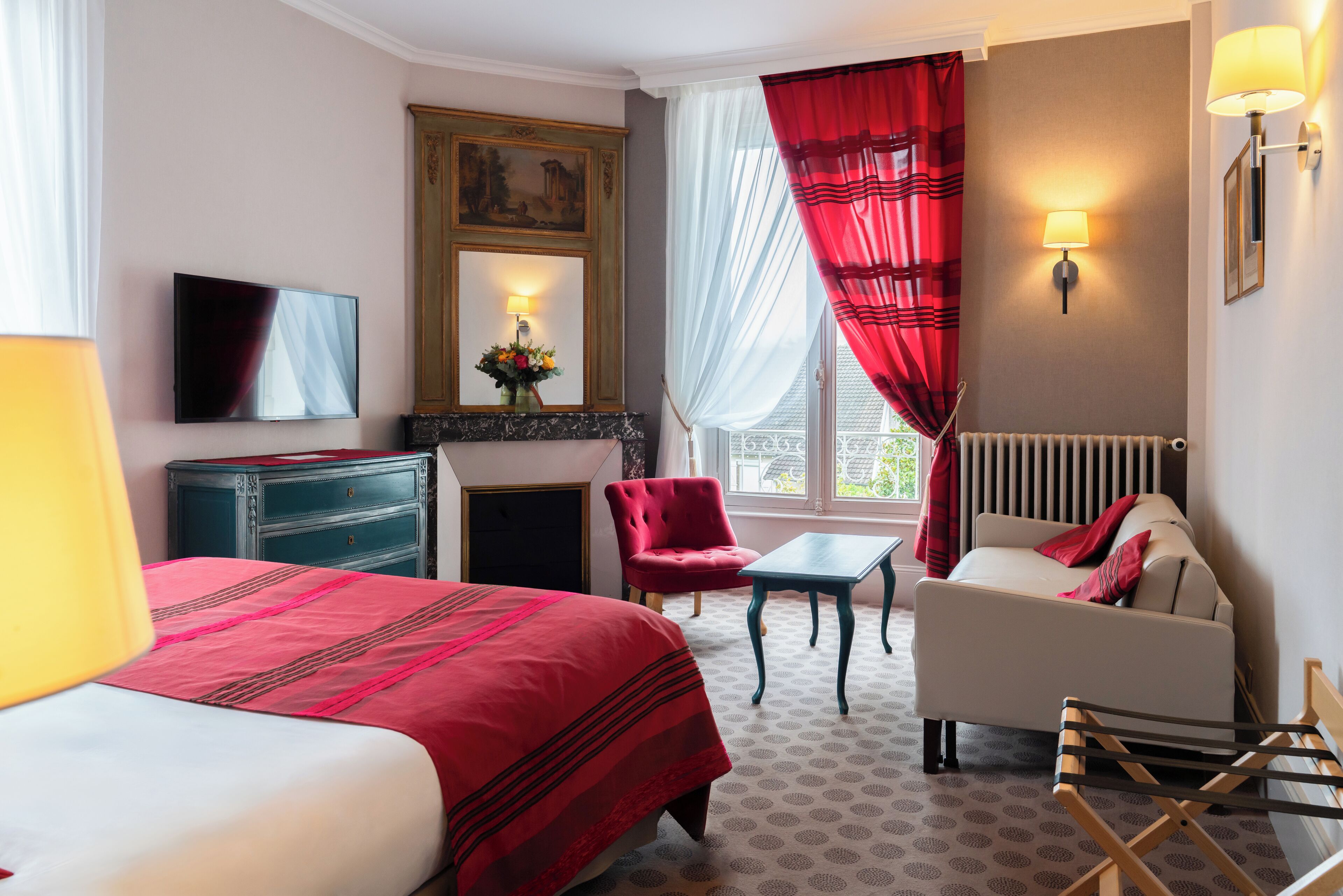 Habitación de hotel con cama, sofá, sillón, mesa, chimenea, televisor y ventana con cortinas.