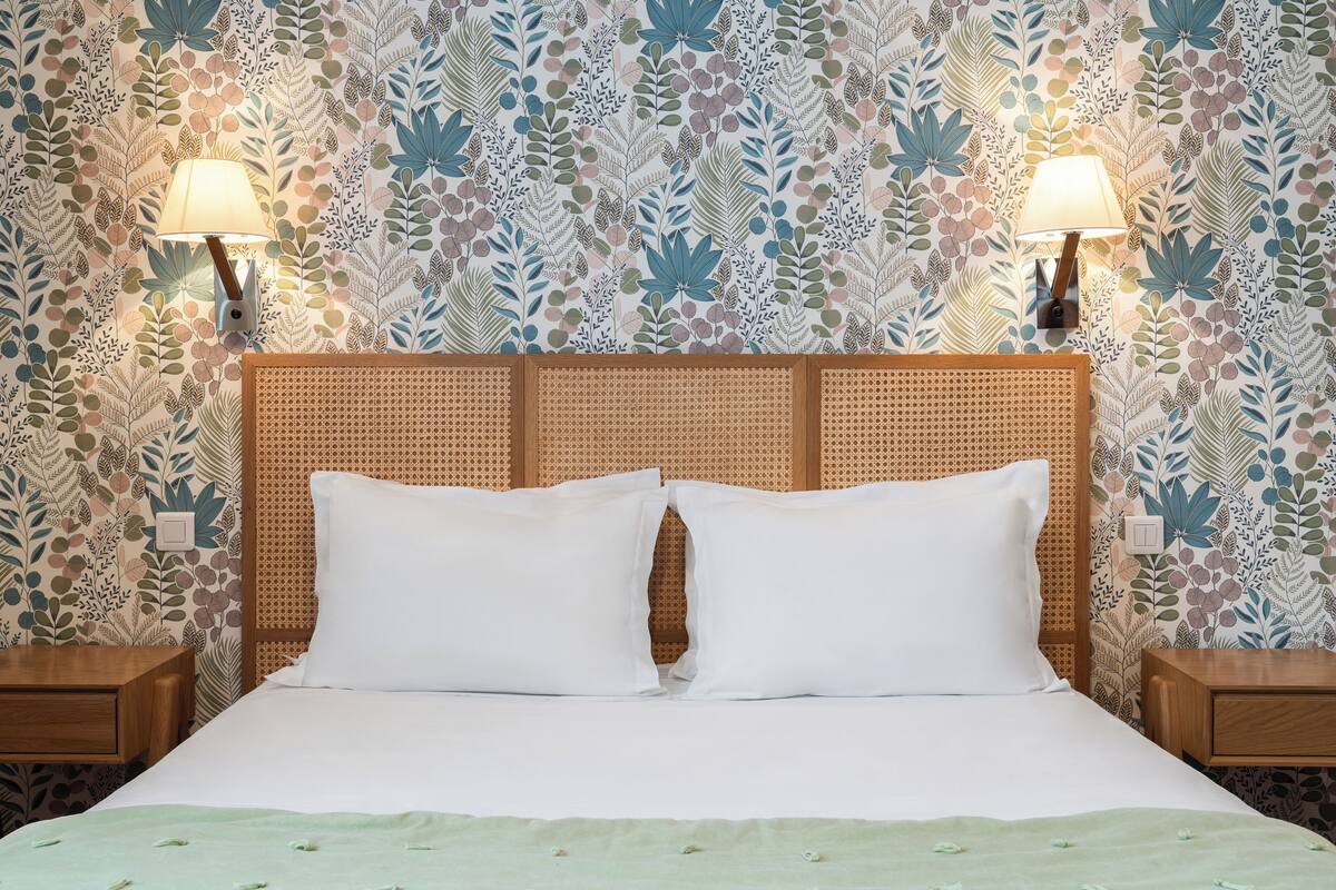 Una cama bien hecha con sábanas blancas, flanqueada por dos mesitas de noche de madera, cada una con una lámpara fijada a la pared. La habitación tiene un papel tapiz estampado con diseño floral.