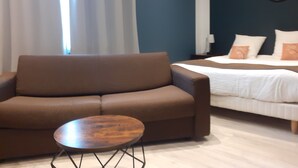 Quarto de hotel moderno com sofá marrom, uma pequena mesa de madeira e uma cama com roupa de cama branca e almofadas decorativas.