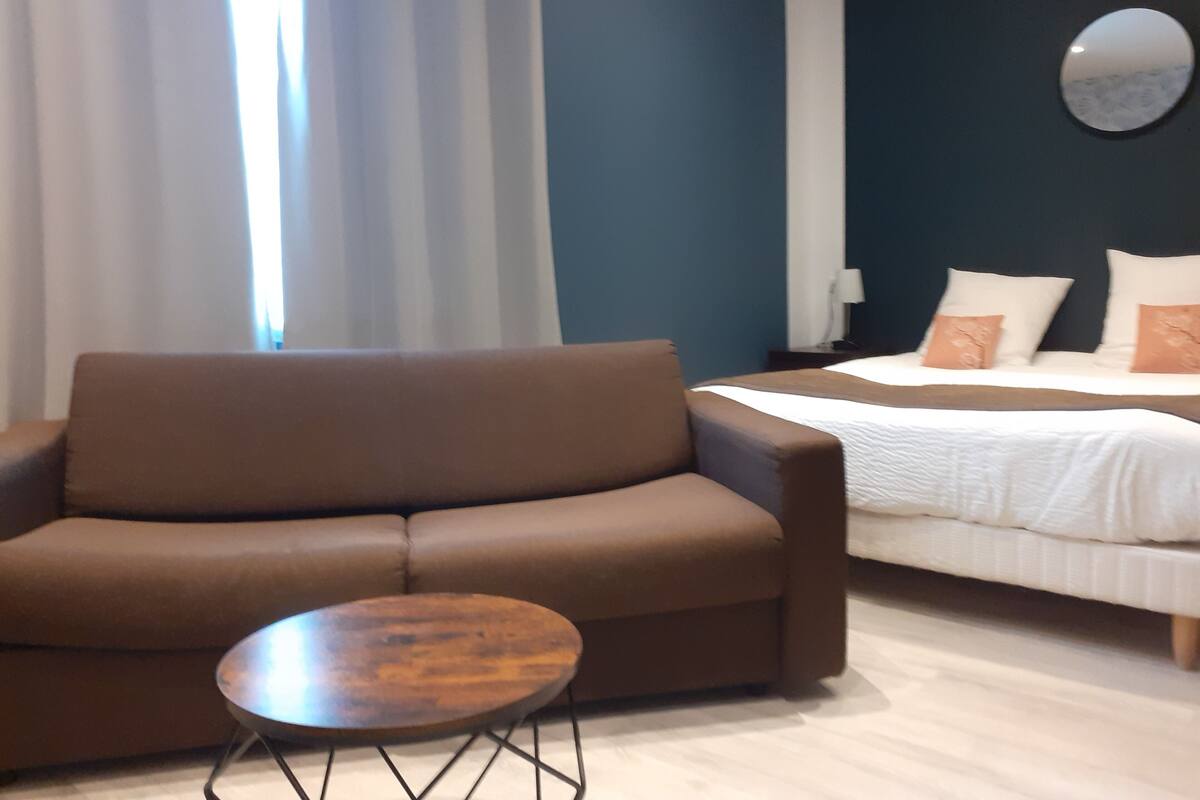 A modern hotel room with a brown sofa, a small wooden table, and a bed with white bedding and decorative pillows.