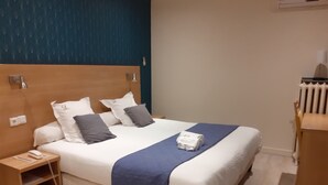 Cama bem arrumada com manta azul, cabeceira de madeira e mesa de cabeceira com um telefone e um pequeno objeto.