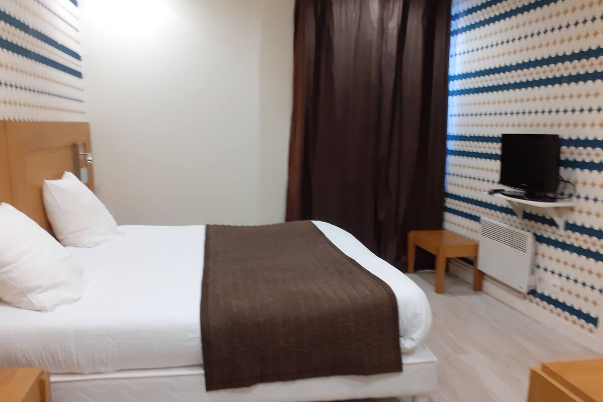 A neatly made bed with a white and brown blanket, a wooden nightstand, a television on a stand, and a patterned wall.