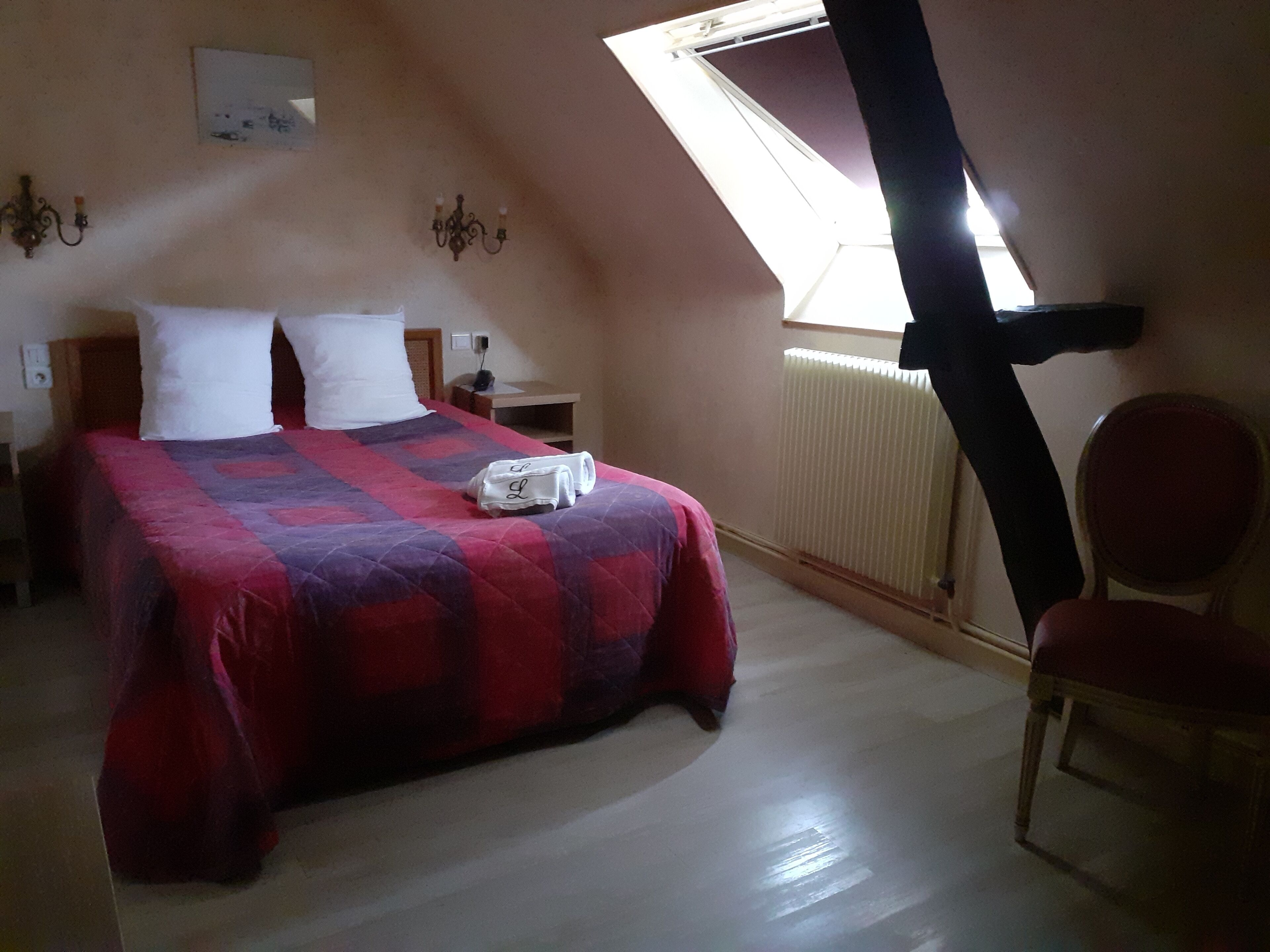 A bedroom with a bed, a chair, a nightstand, a lamp, a radiator, and a window.