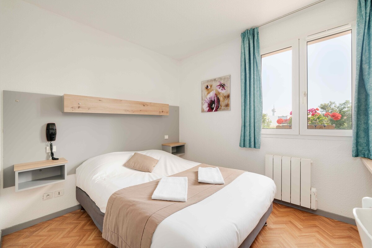 A hotel room with a large bed, a bedside table, a wall-mounted phone, a radiator, and a window with curtains.