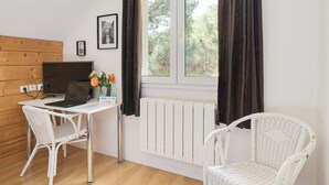 Una habitación con pared de madera, un radiador blanco, un escritorio con una computadora portátil, dos sillas blancas de mimbre y una ventana con cortinas.