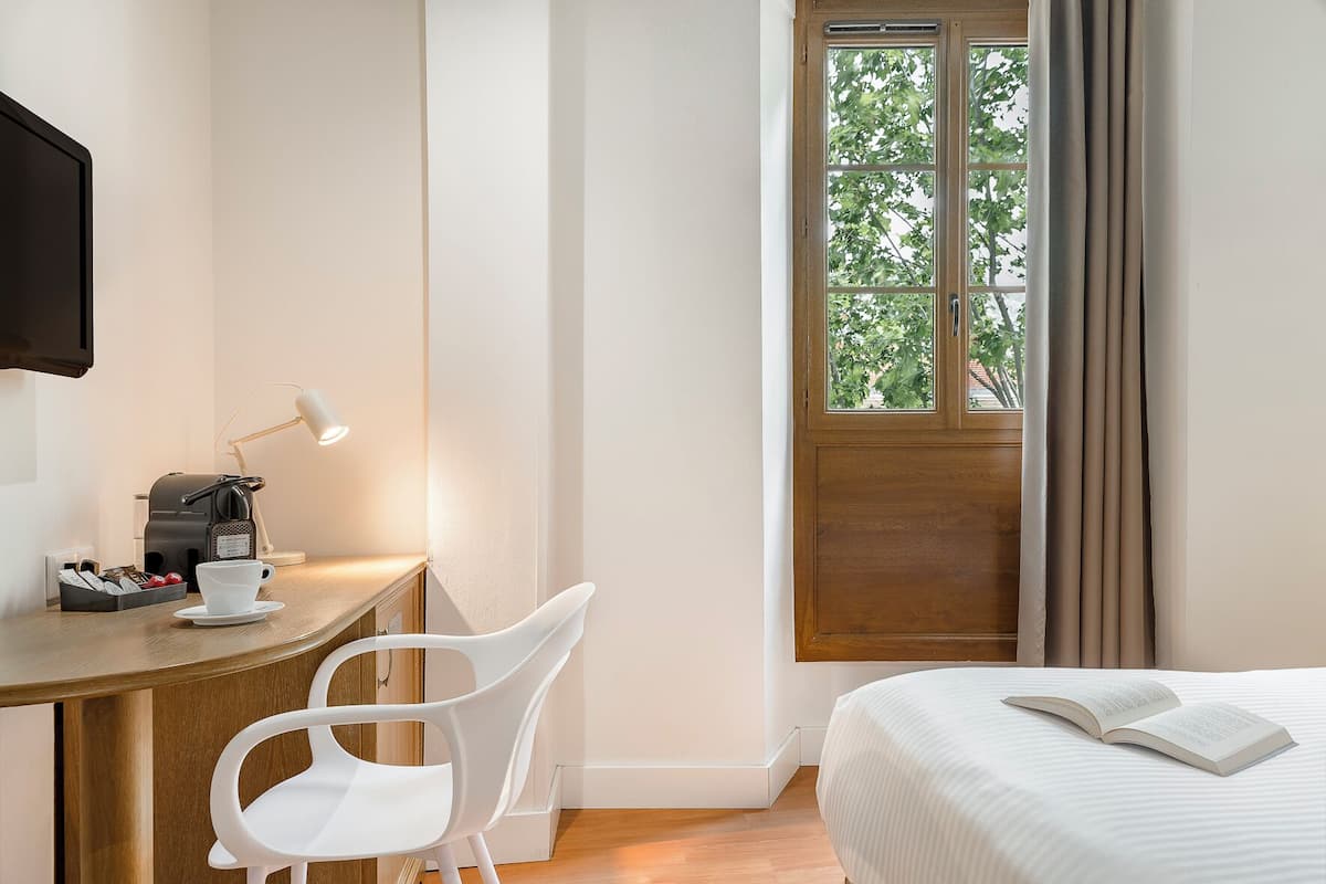 A hotel room with a wooden desk, a white chair, a bed with a white comforter, a window with curtains, and a television mounted on the wall.