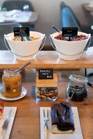 Dos tazones de cereal con etiquetas, un tarro de miel, un recipiente con frutos secos y un postre de chocolate sobre una mesa de madera.