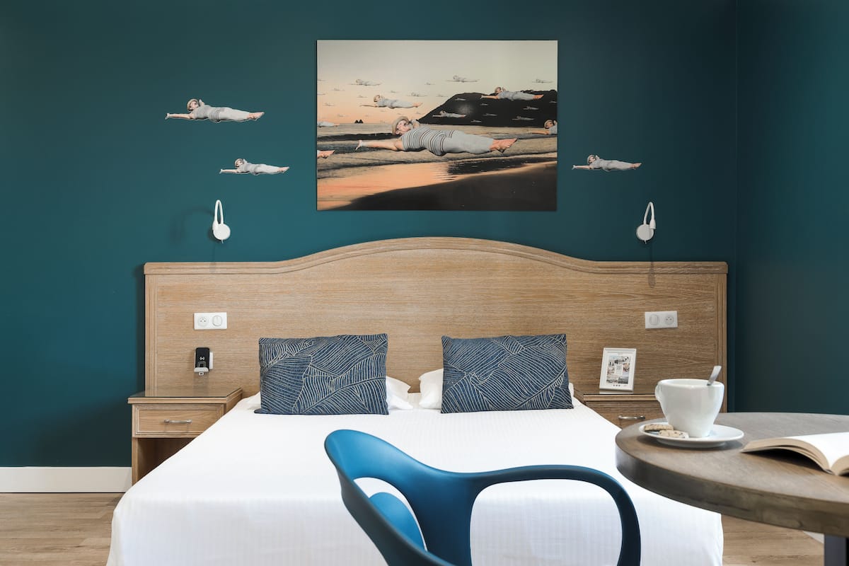 A hotel room with a bed, a bedside table, a chair, a small table with a cup and book, and a wall with a painting.