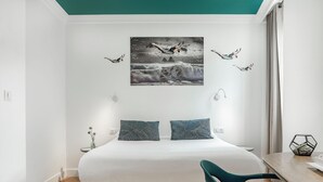 Une chambre moderne avec un grand lit, deux oreillers, une table de chevet, une chaise et une œuvre d’art fixée au mur représentant des oiseaux en vol.