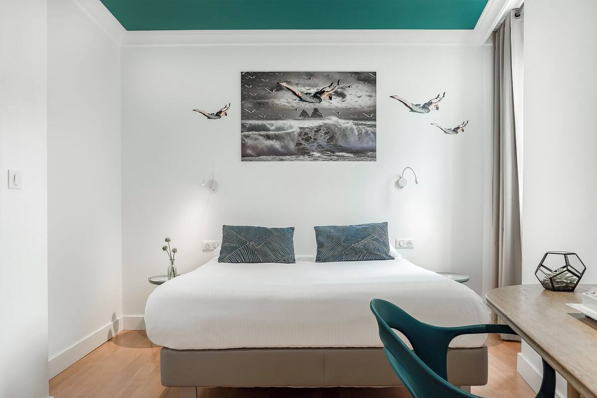 A modern bedroom with a large bed, two pillows, a side table, a chair, and a wall-mounted artwork depicting birds in flight.