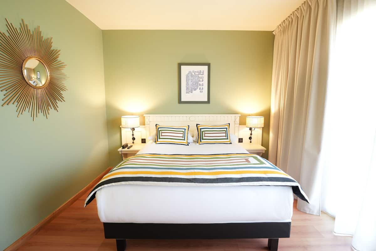 A hotel room with a bed, two bedside lamps, a framed picture on the wall, and a sunburst mirror on the left.