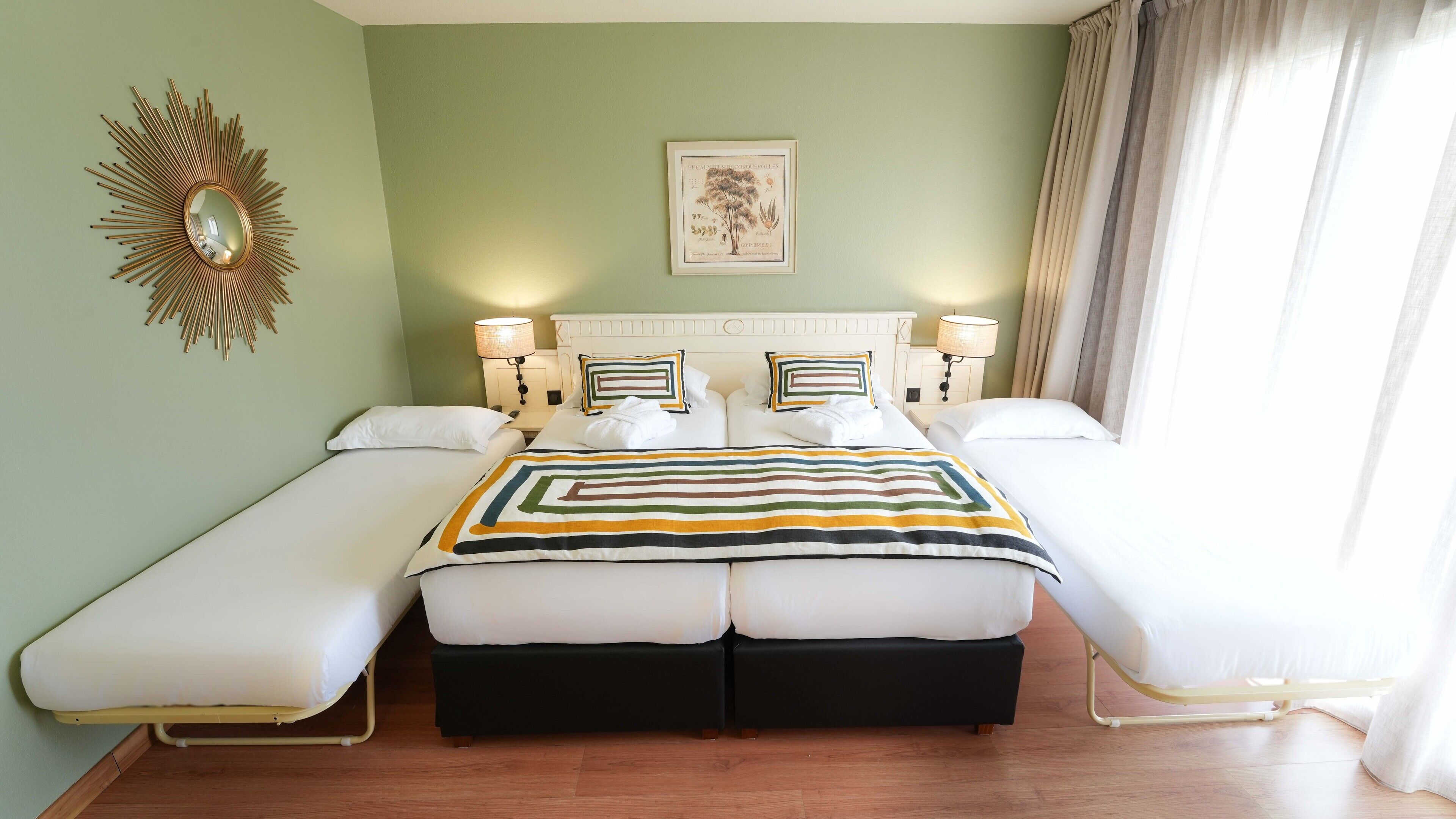 Un lit double avec une couvre-lit à rayures, deux tables de chevet avec des lampes, un miroir fixé au mur et une illustration botanique encadrée.