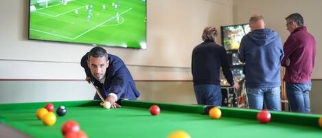 A pool table with a person playing, a TV displaying a soccer match, and three people watching.