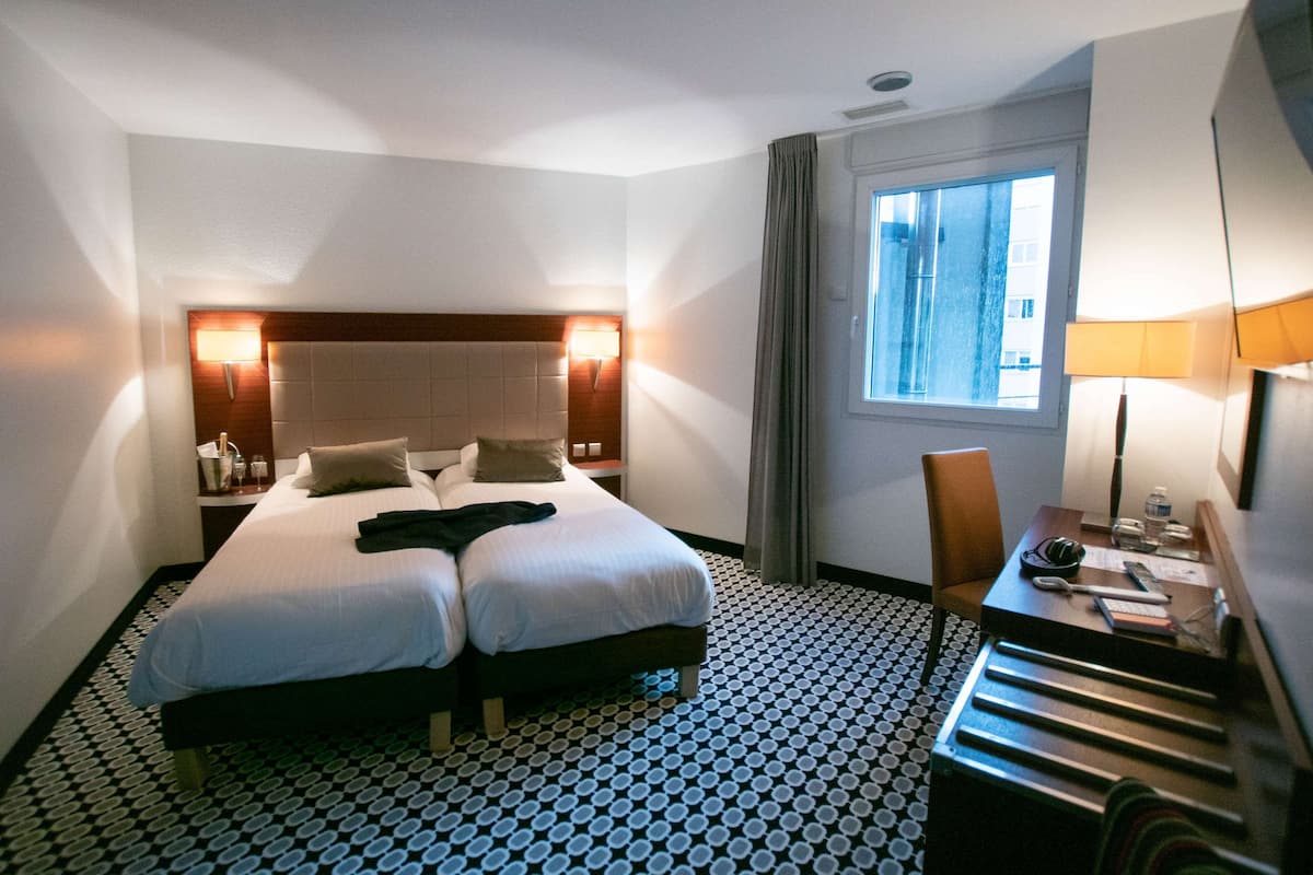 A hotel room with two beds, a desk, a chair, a window, and a patterned floor.