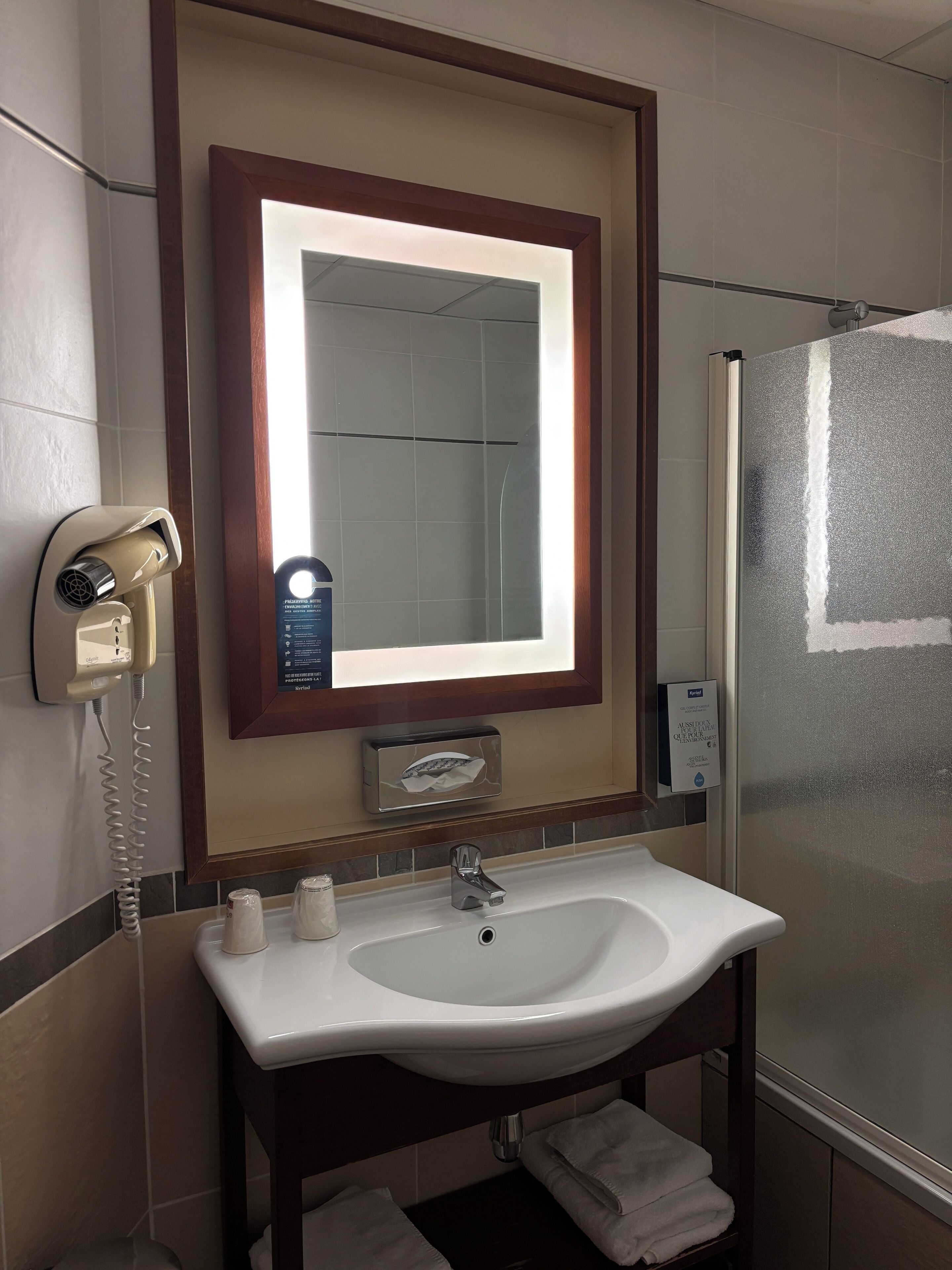 A bathroom with a white sink, a mirror with a light, a hairdryer, and a glass shower enclosure.