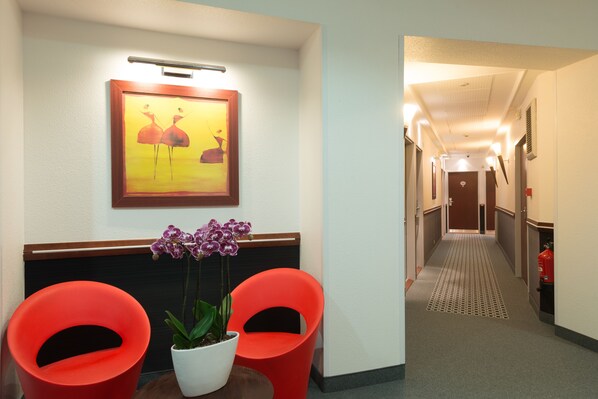 A hotel lobby with a reception desk, two red chairs, a potted orchid, and a framed artwork on the wall.