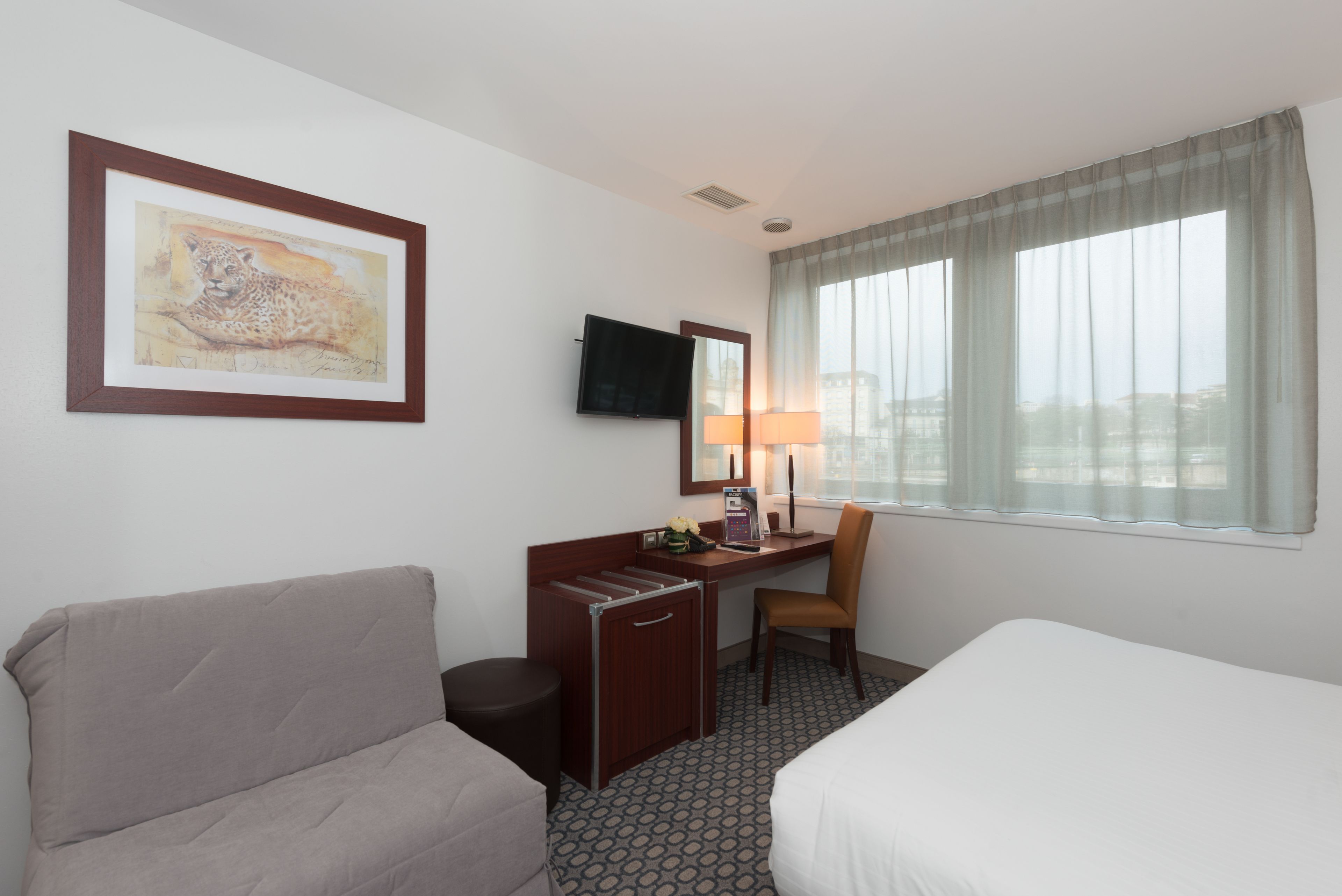 Habitación de hotel con cama, escritorio y silla, televisor, un cuadro en la pared y ventana con cortinas.