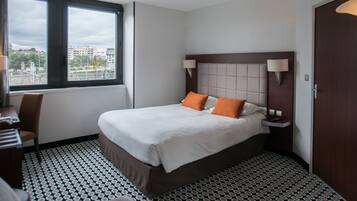 A hotel room with a bed, two bedside lamps, a desk with a chair, a window with a view of buildings, and a patterned floor.