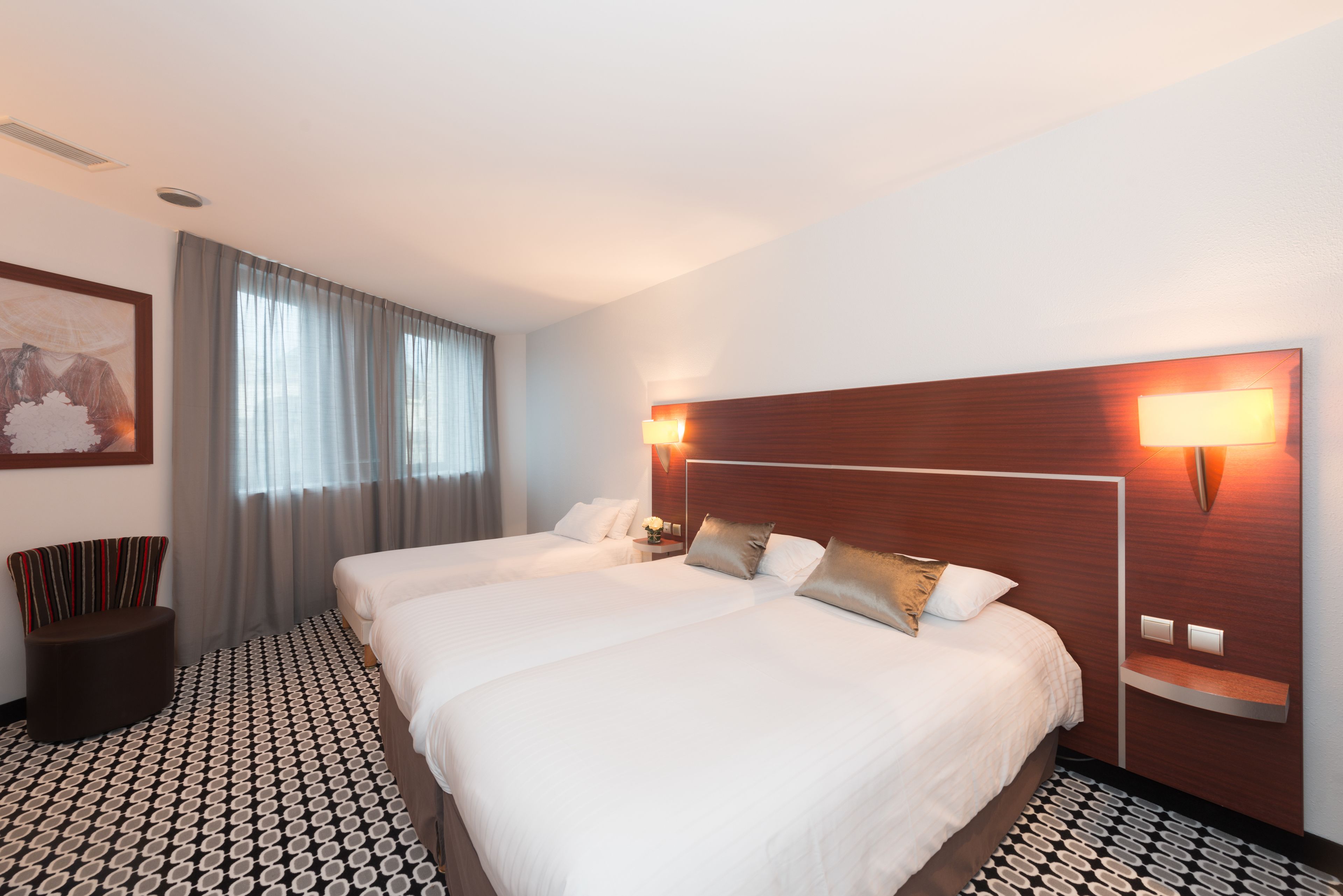 Habitación de hotel con dos camas, un ventanal con cortinas, una lámpara de pared y un suelo estampado.