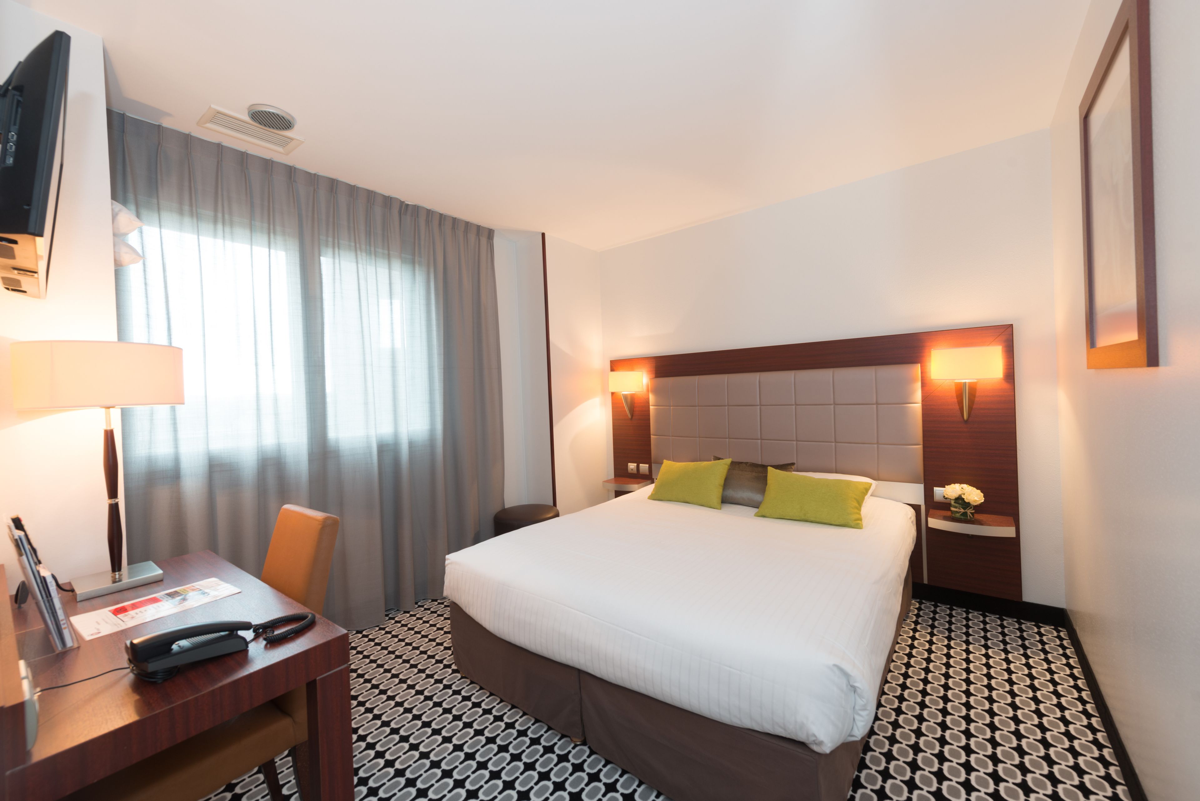 A hotel room with a large bed, a desk with a telephone, a chair, a TV, and a window with curtains.
