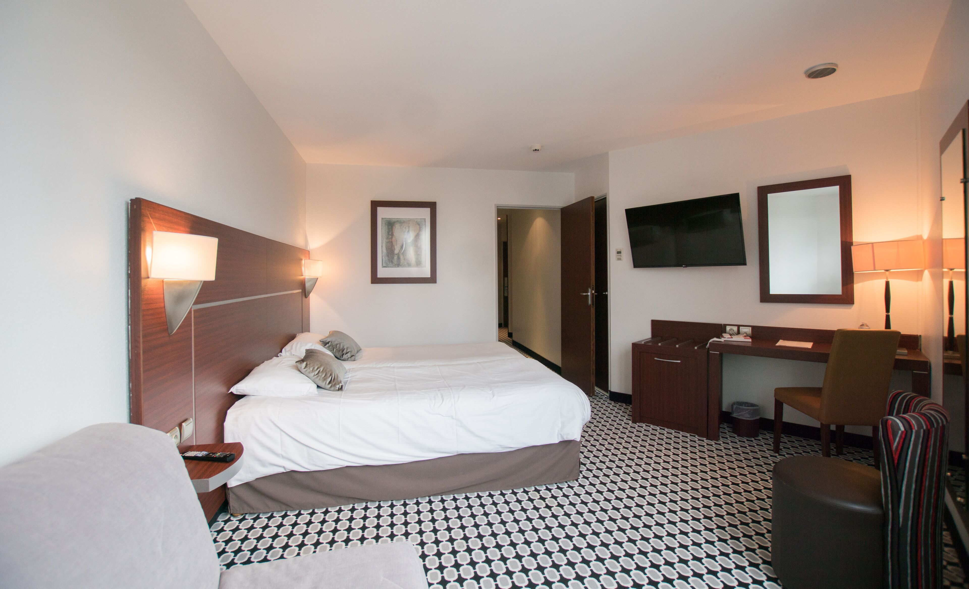 A hotel room with a bed, bedside tables, a desk, a chair, a television, and a mirror.