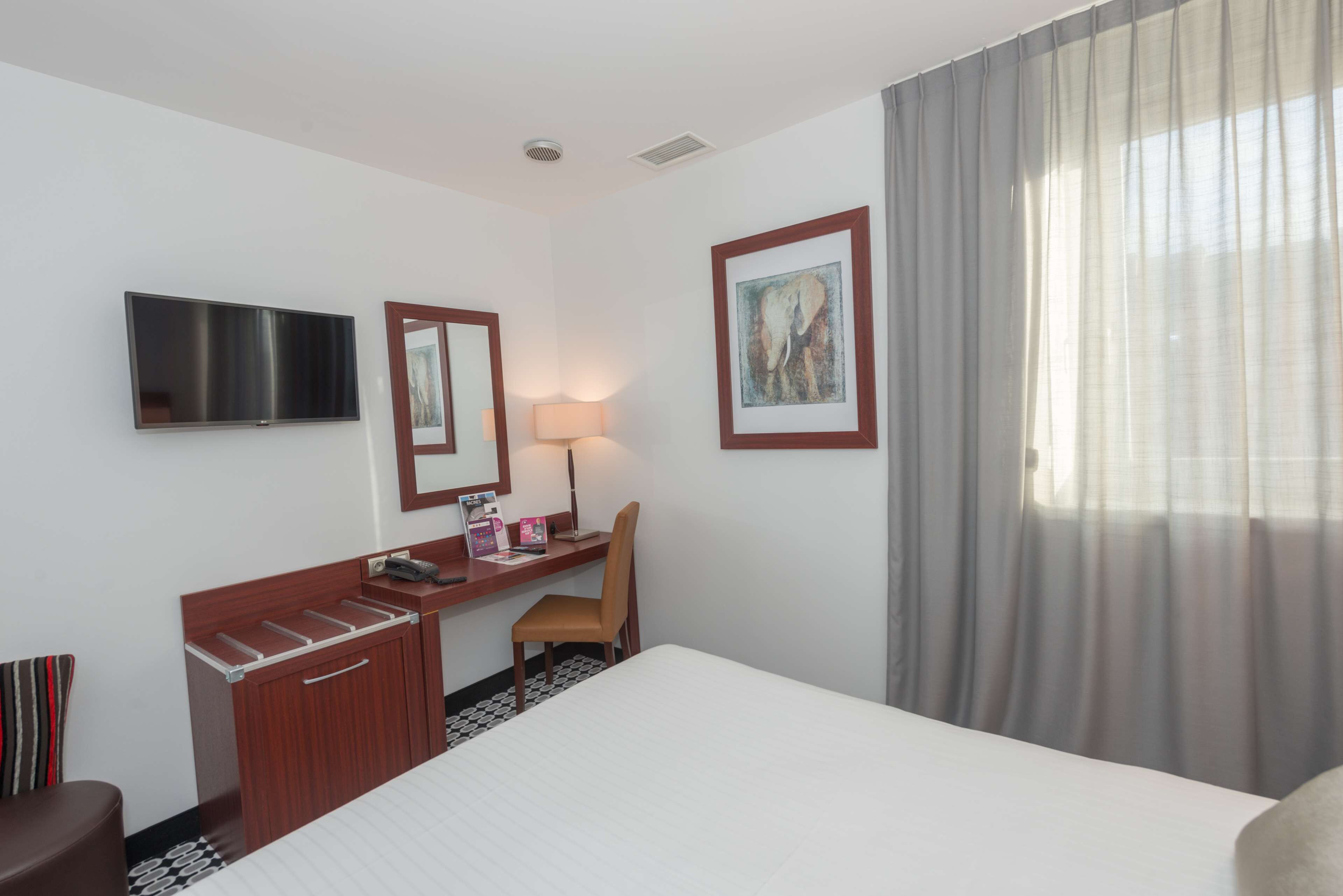 A hotel room with a bed, a desk with a chair, a TV, a lamp, and a framed picture on the wall.