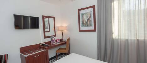 A hotel room with a bed, a desk with a chair, a TV, a lamp, and a framed picture on the wall.