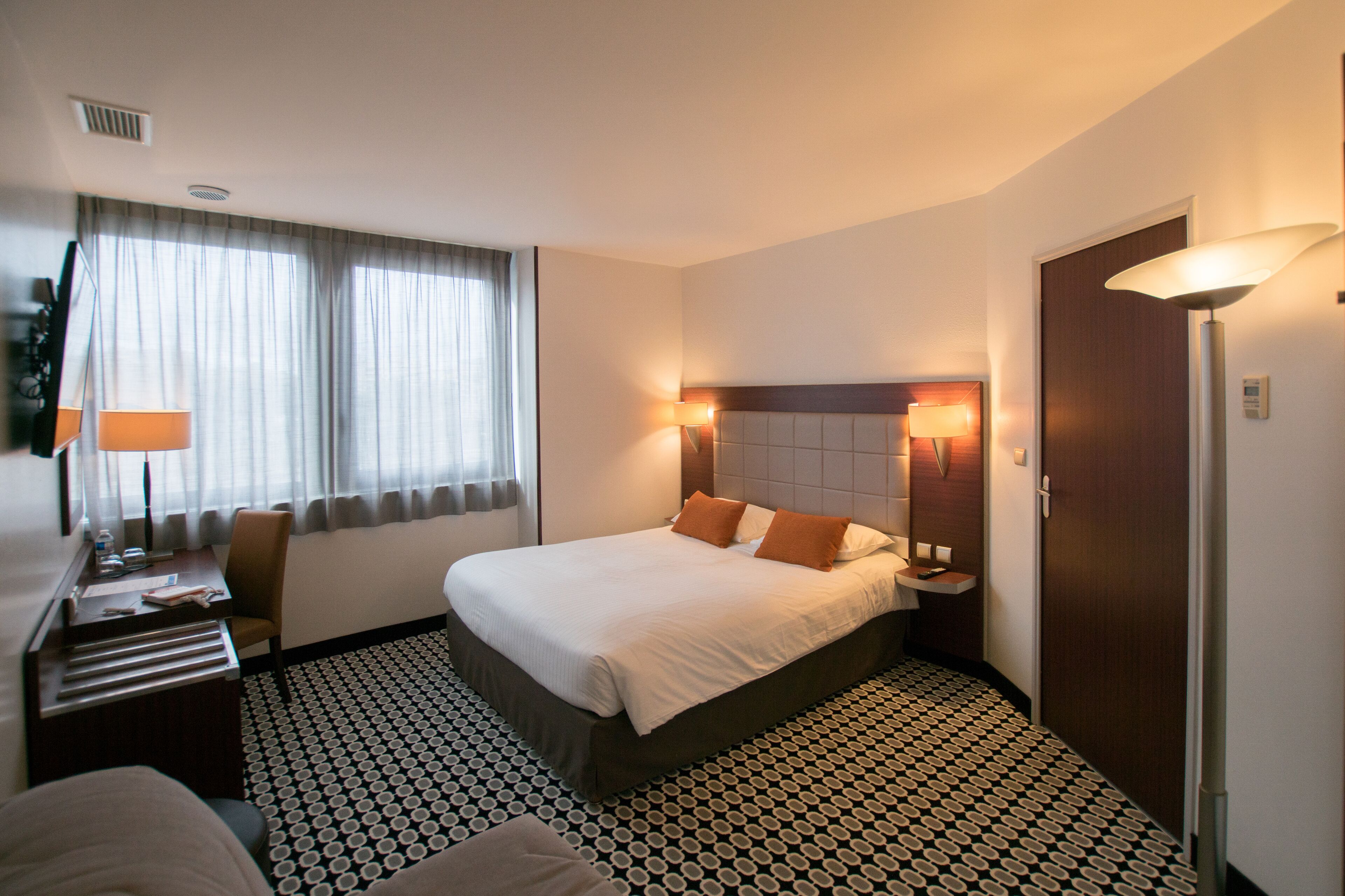 A hotel room with a bed, bedside lamps, a desk, and a patterned floor.