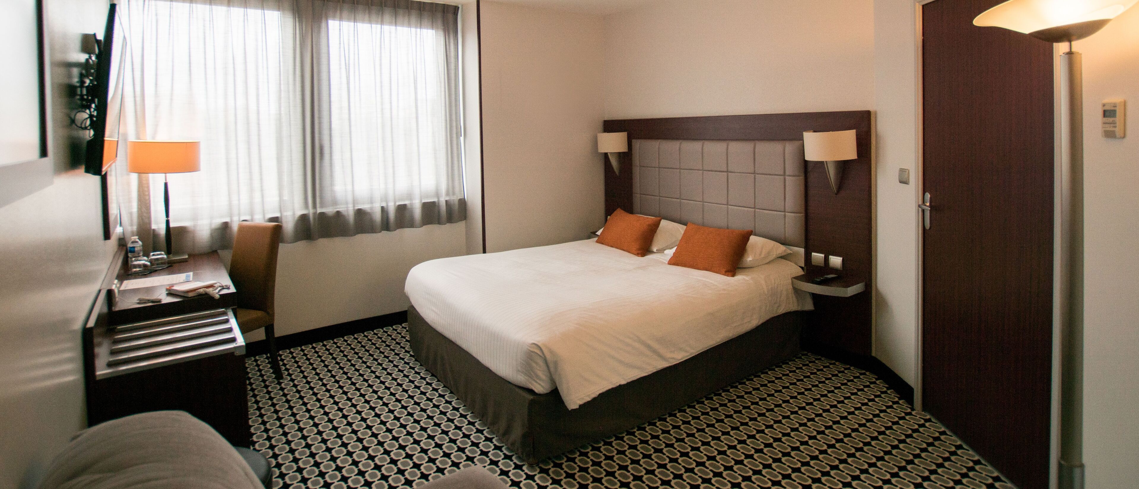 A hotel room with a bed, bedside lamps, a desk with a lamp, and a patterned floor.