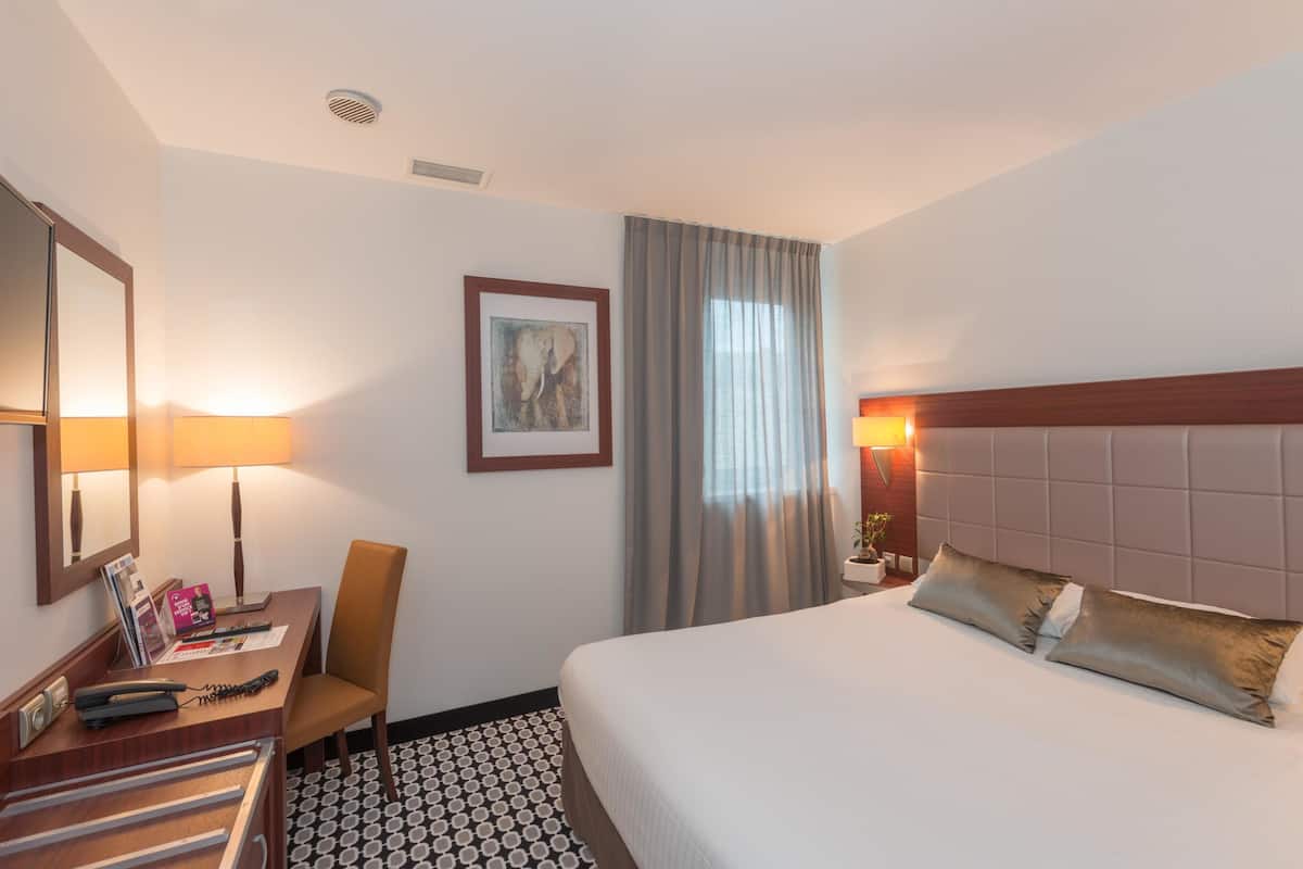 A hotel room with a bed, a desk with a lamp, a chair, a framed picture on the wall, and a window with curtains.
