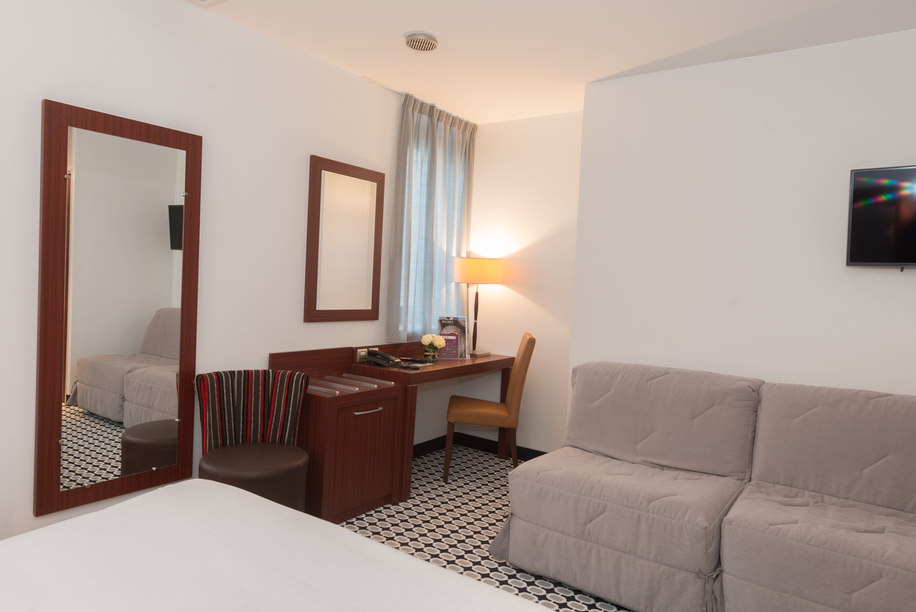 A hotel room with a bed, a desk with a chair, a sofa, a mirror, a lamp, and a television.