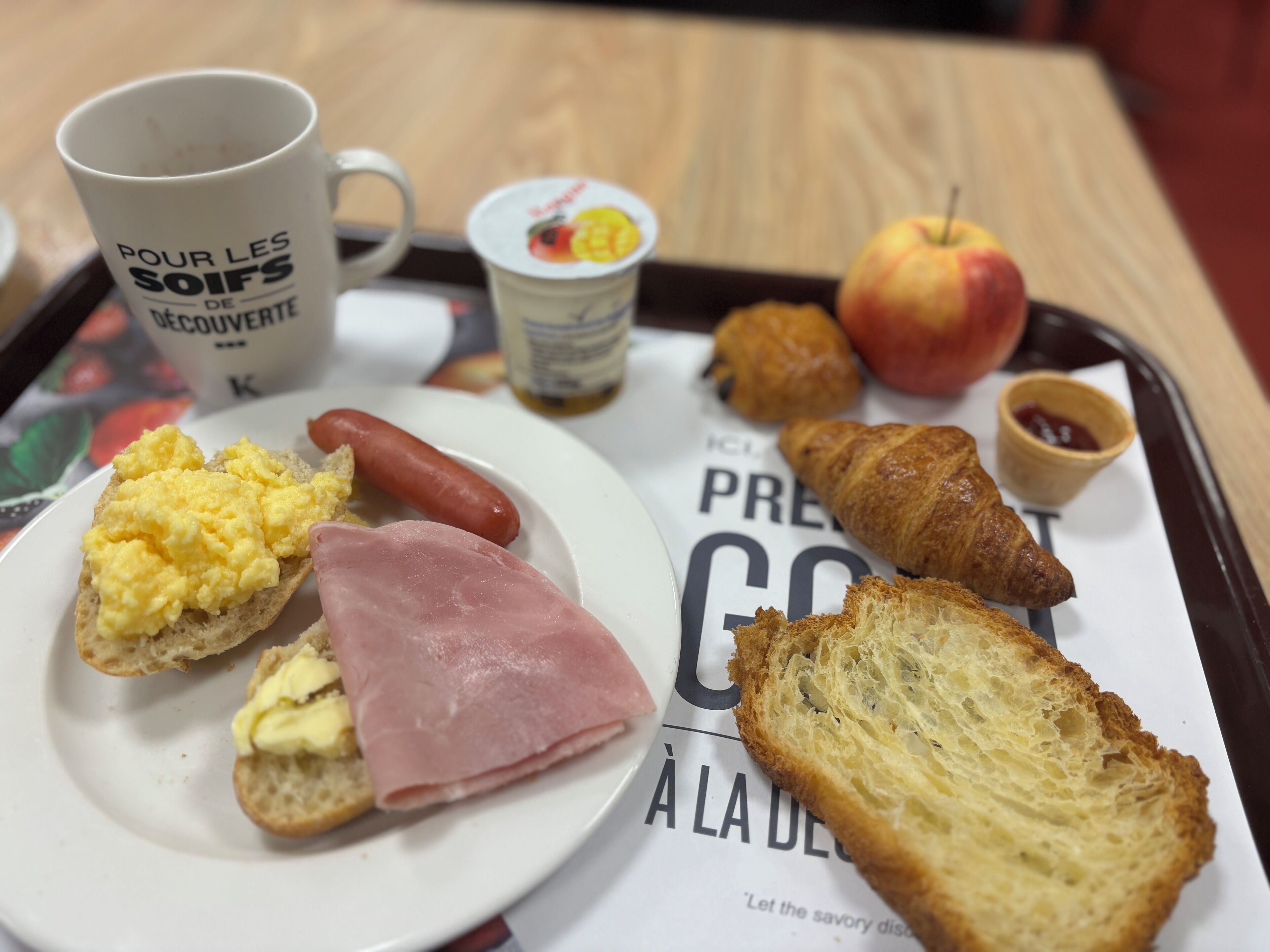 Bandeja de desayuno con huevos revueltos, jamón, salchicha, croissant, manzana y una taza de café.