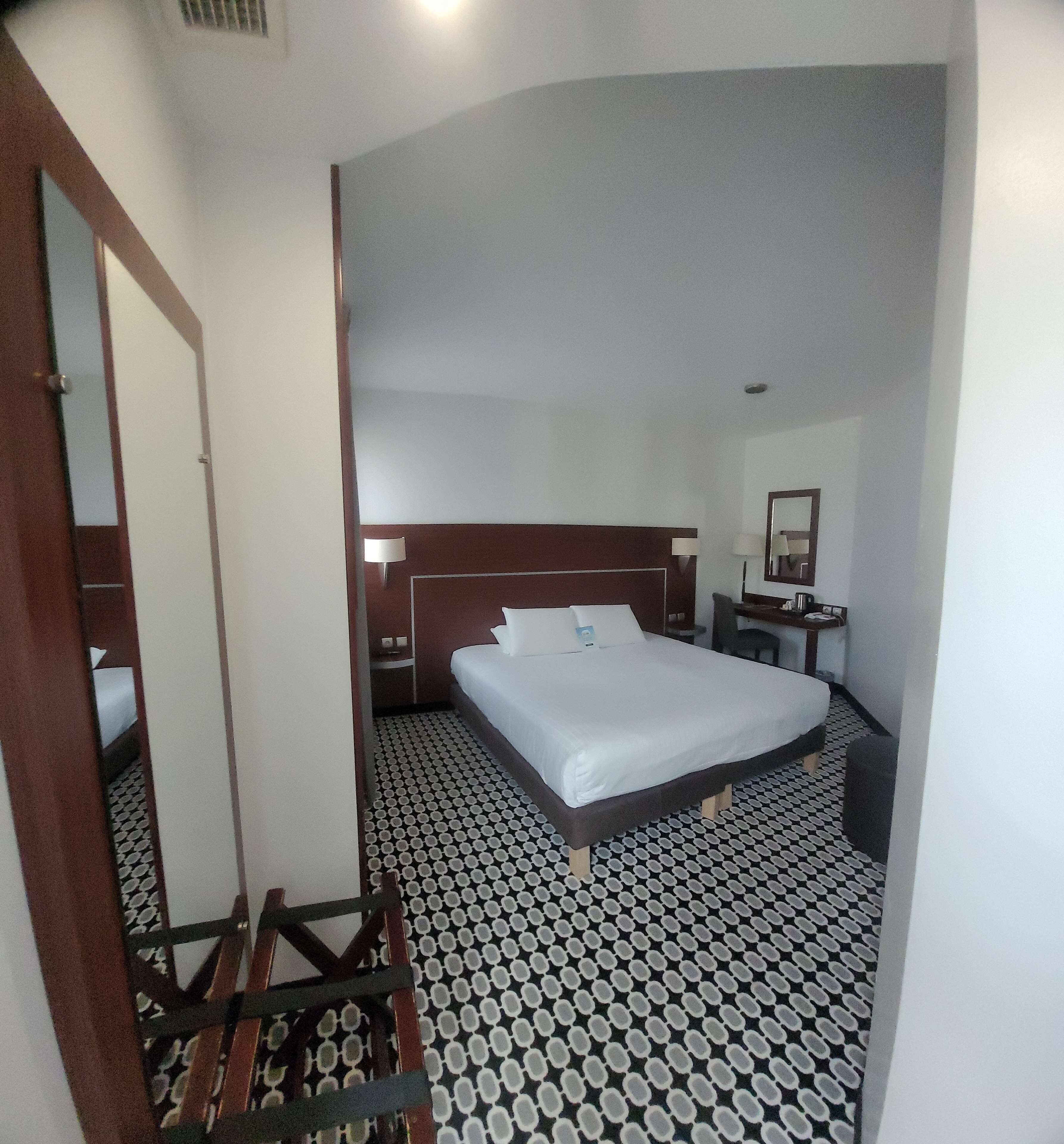 Une chambre d’hôtel avec un lit, un bureau, une chaise, un miroir et un sol à motifs.
