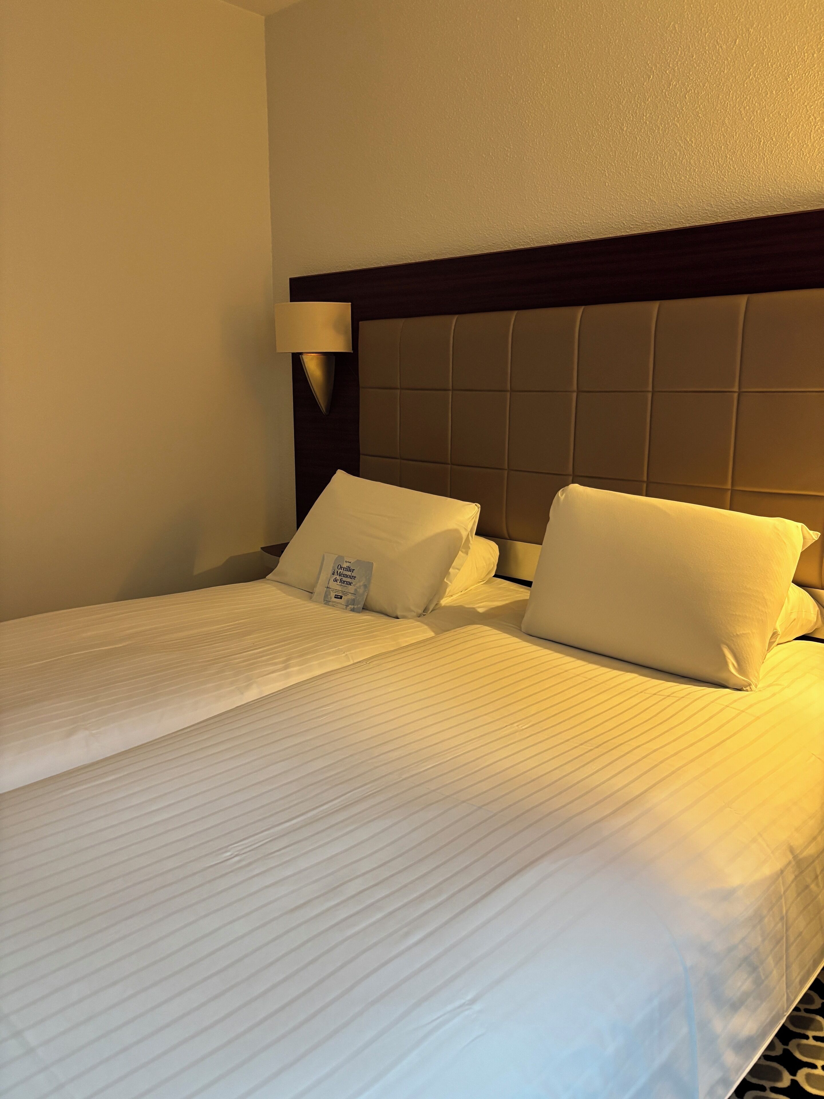 Une chambre d’hôtel avec deux lits, chacun doté de draps et d’oreillers blancs, d’une tête de lit sombre et d’une lampe de chevet.