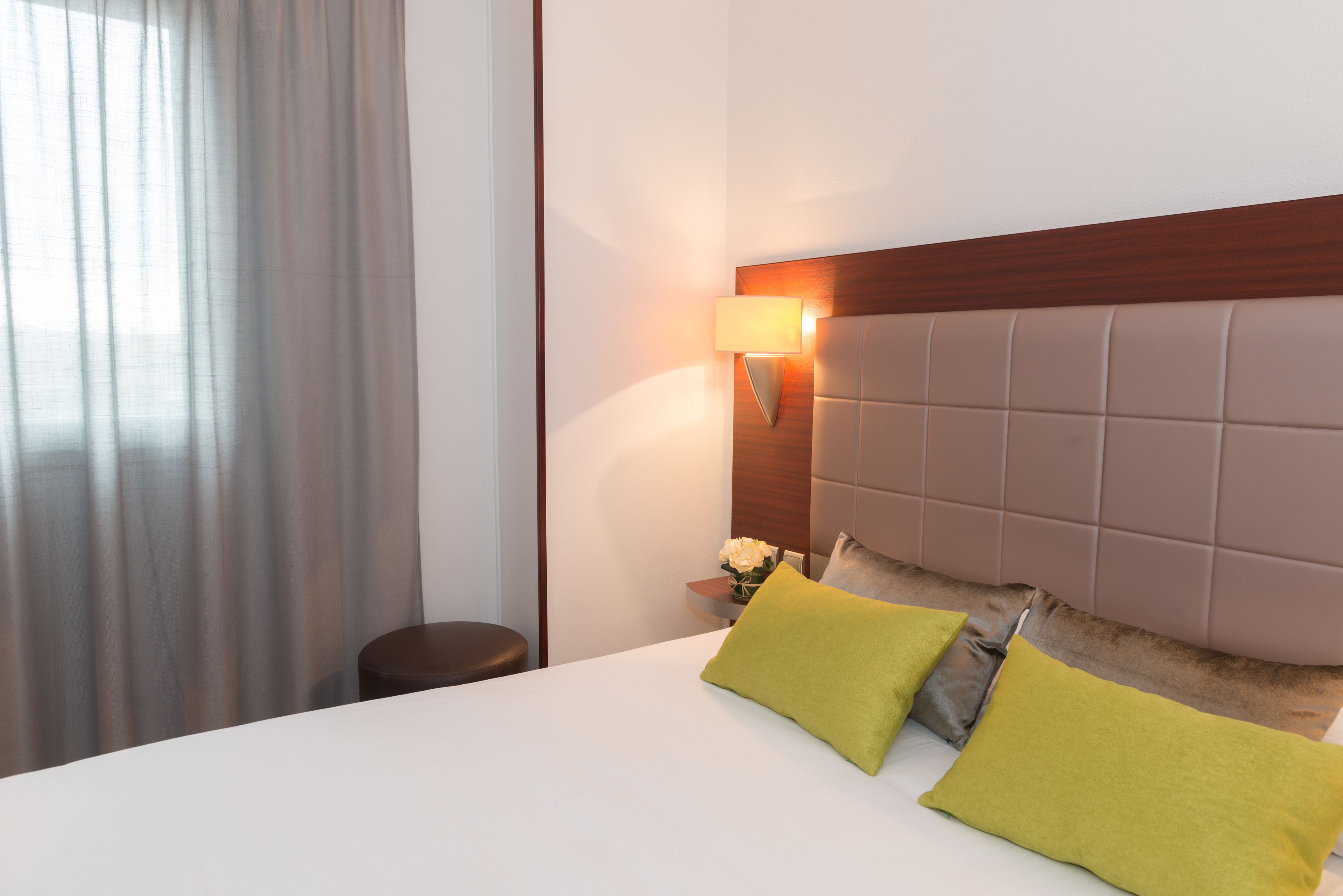 A hotel room with a large bed, a wooden headboard, a nightstand with a lamp, and a window with curtains.