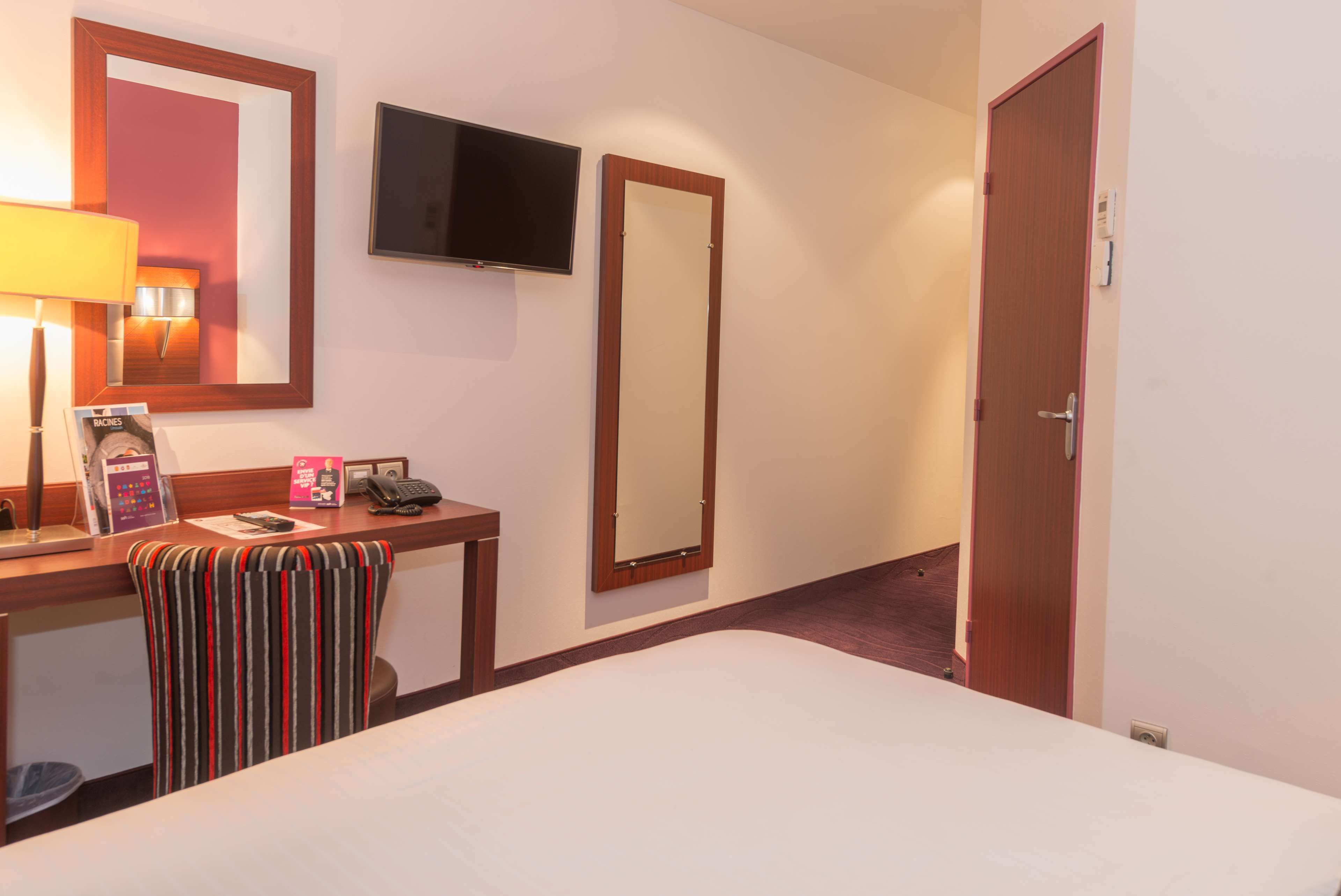 A hotel room with a bed, a desk with a telephone, a mirror, and a television mounted on the wall.