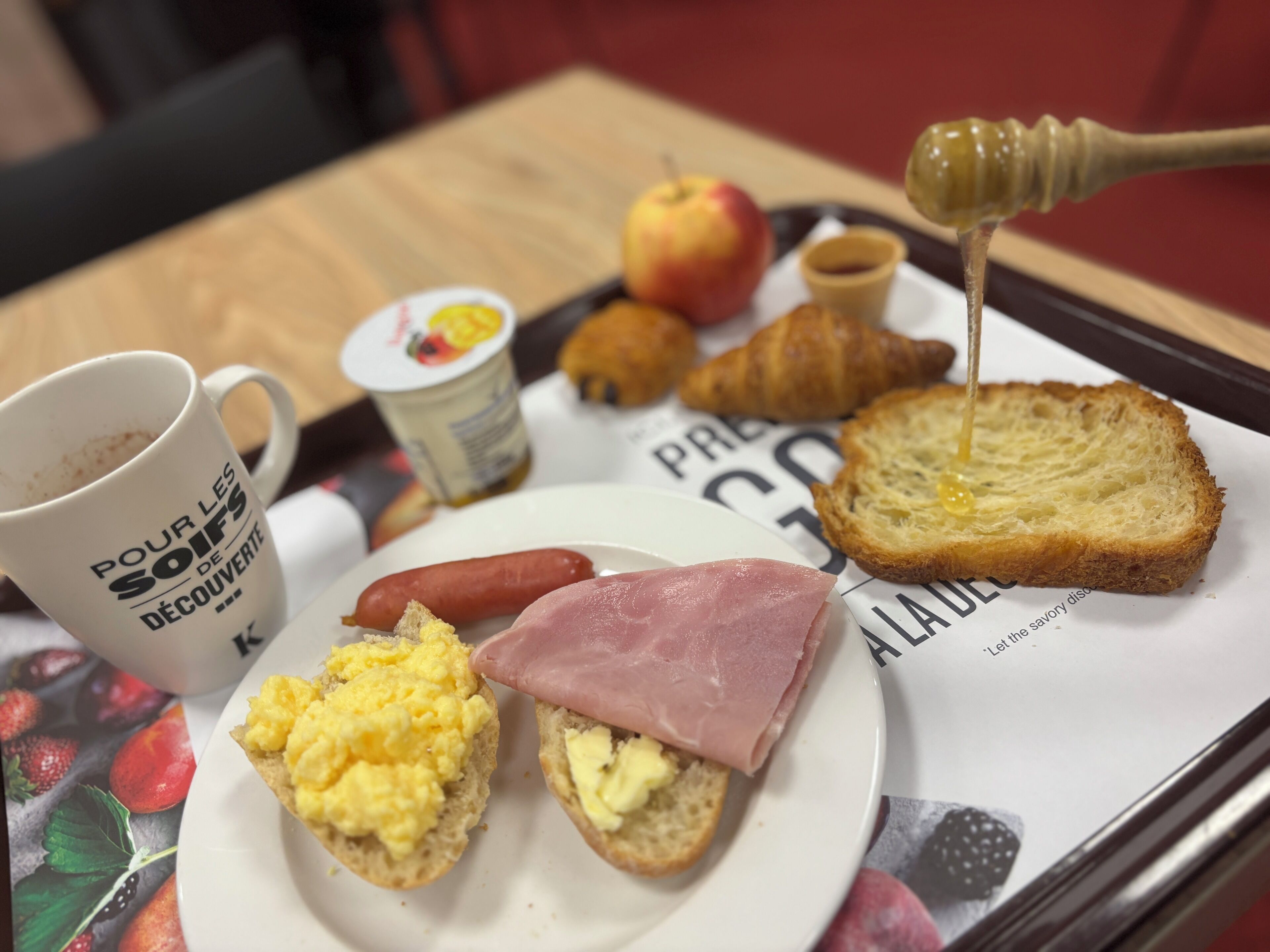 Bandeja de desayuno con tostadas, huevos revueltos, jamón, salchichas, un croissant, una taza de café y un gotero de miel.