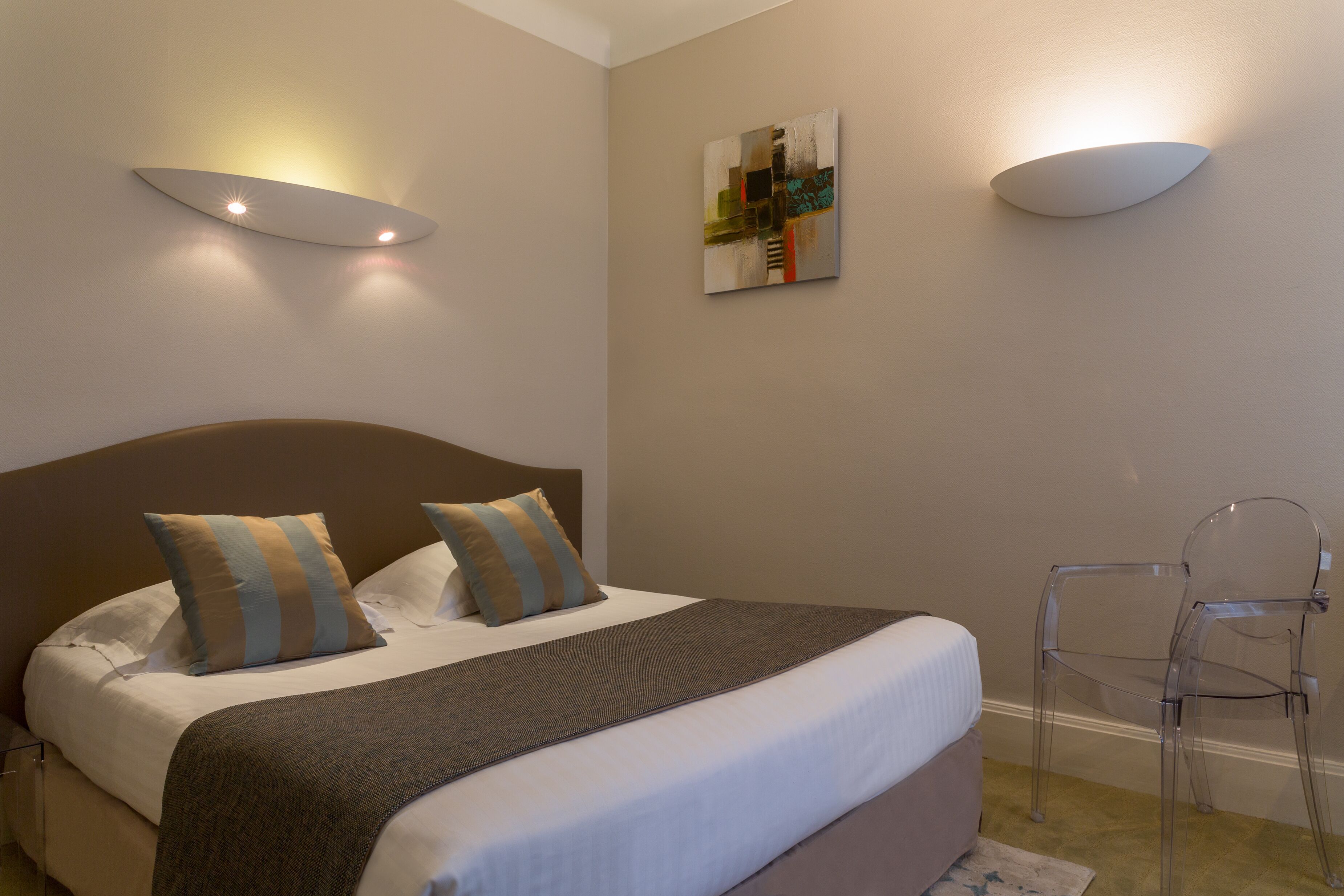 Una cama bien hecha con cabecera marrón, dos almohadas y una manta gris. Un sillón de plástico transparente a la derecha y una lámpara fijada a la pared sobre la cama.